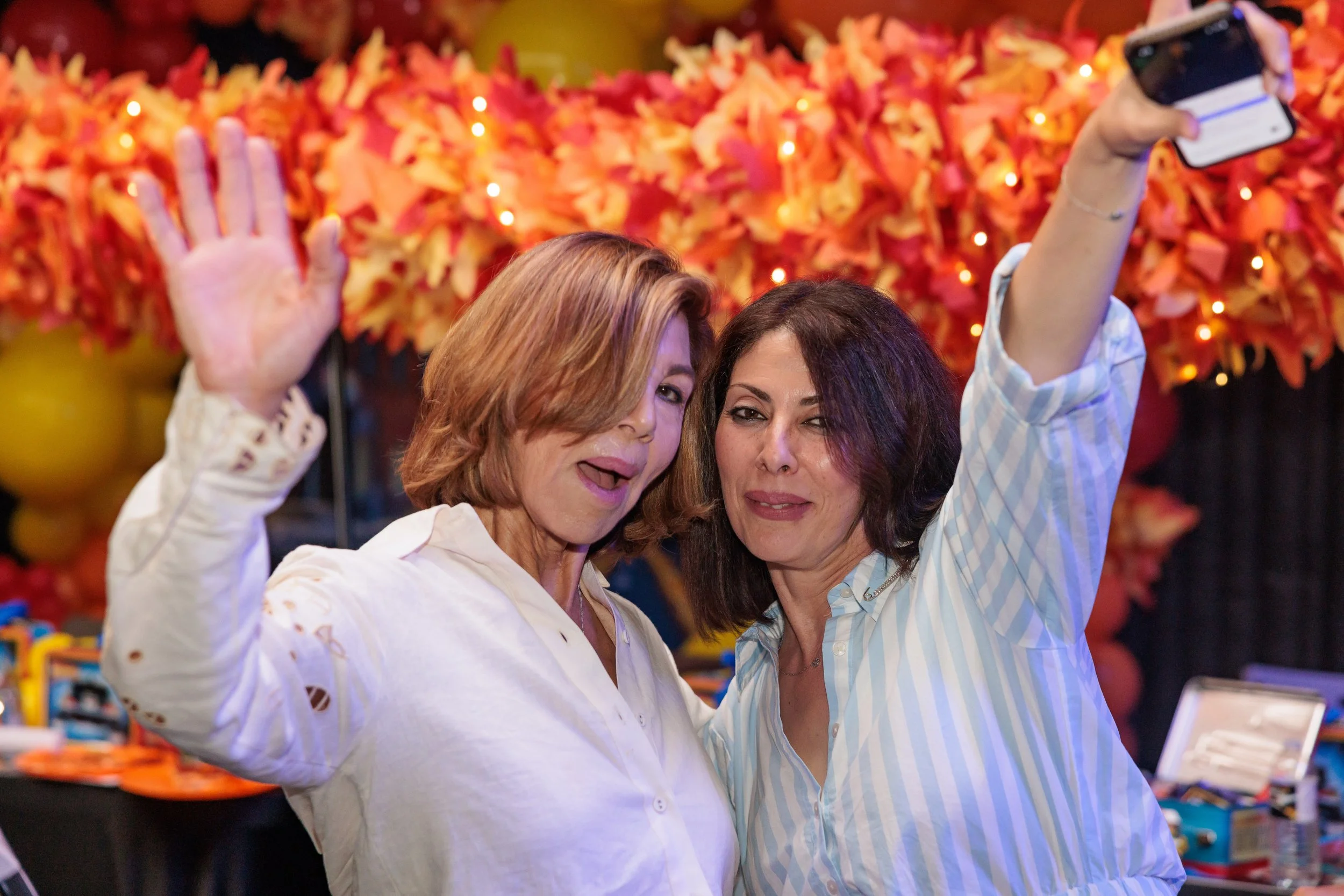 Two women taking a selfie at a festive event with orange and yellow decor in the background.