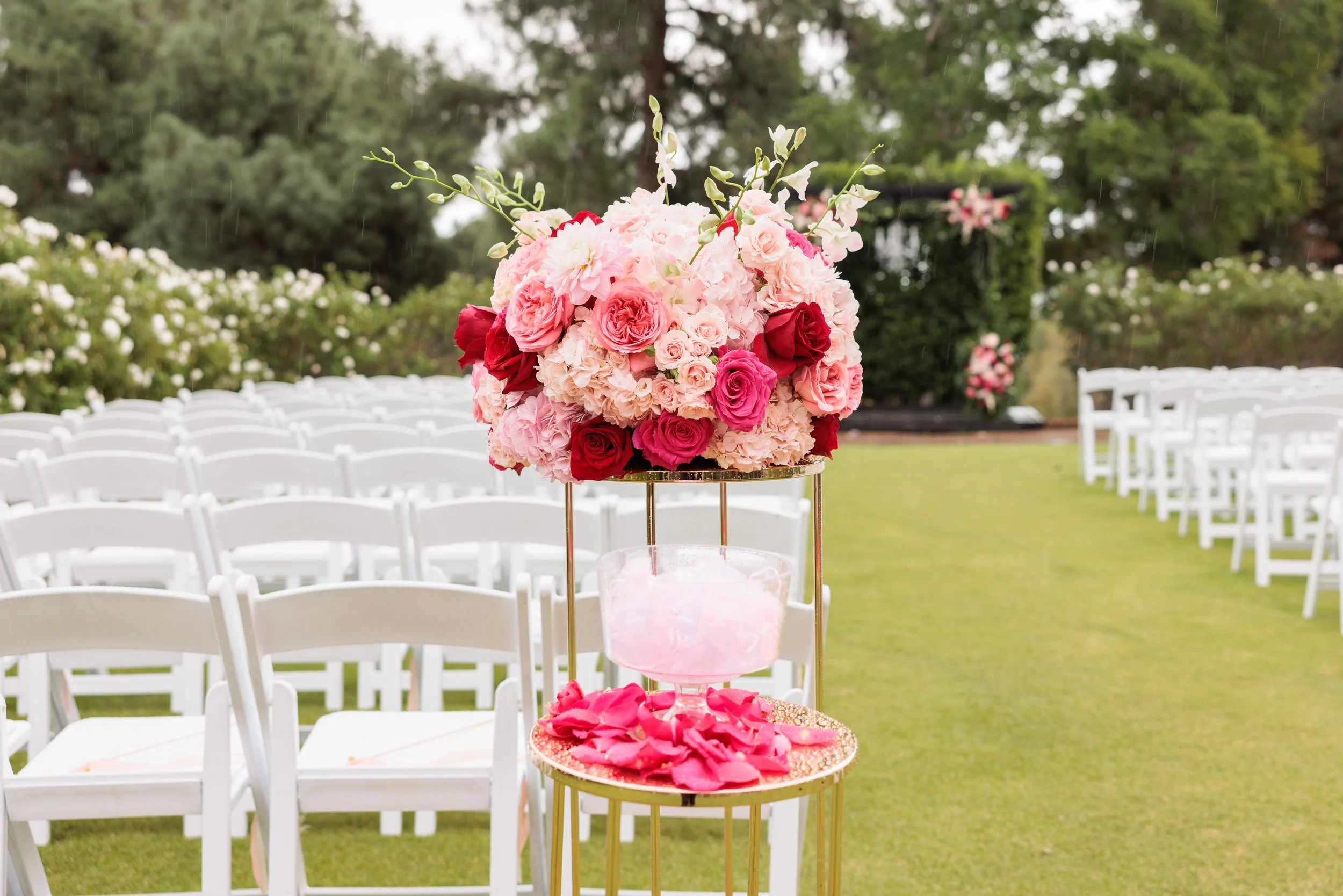 A floral wedding centerpiece with pink, red, and white flowers on a gold stand, set in an outdoor venue with white chairs and greenery.