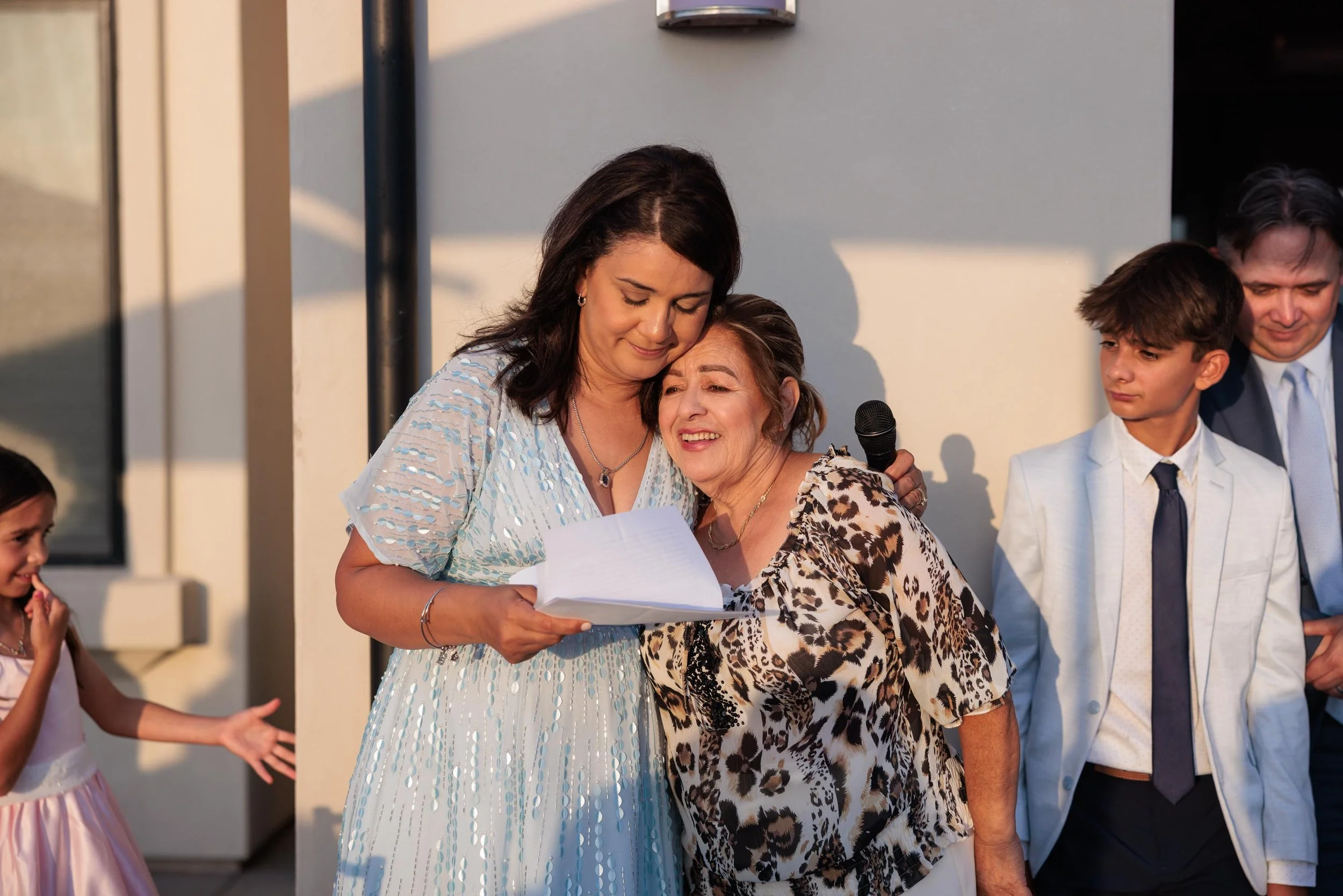 A woman comforting an elderly woman who is crying, while holding a microphone and reading from a paper during an outdoor event. Other people, including children and a man in a suit, are present nearby.
