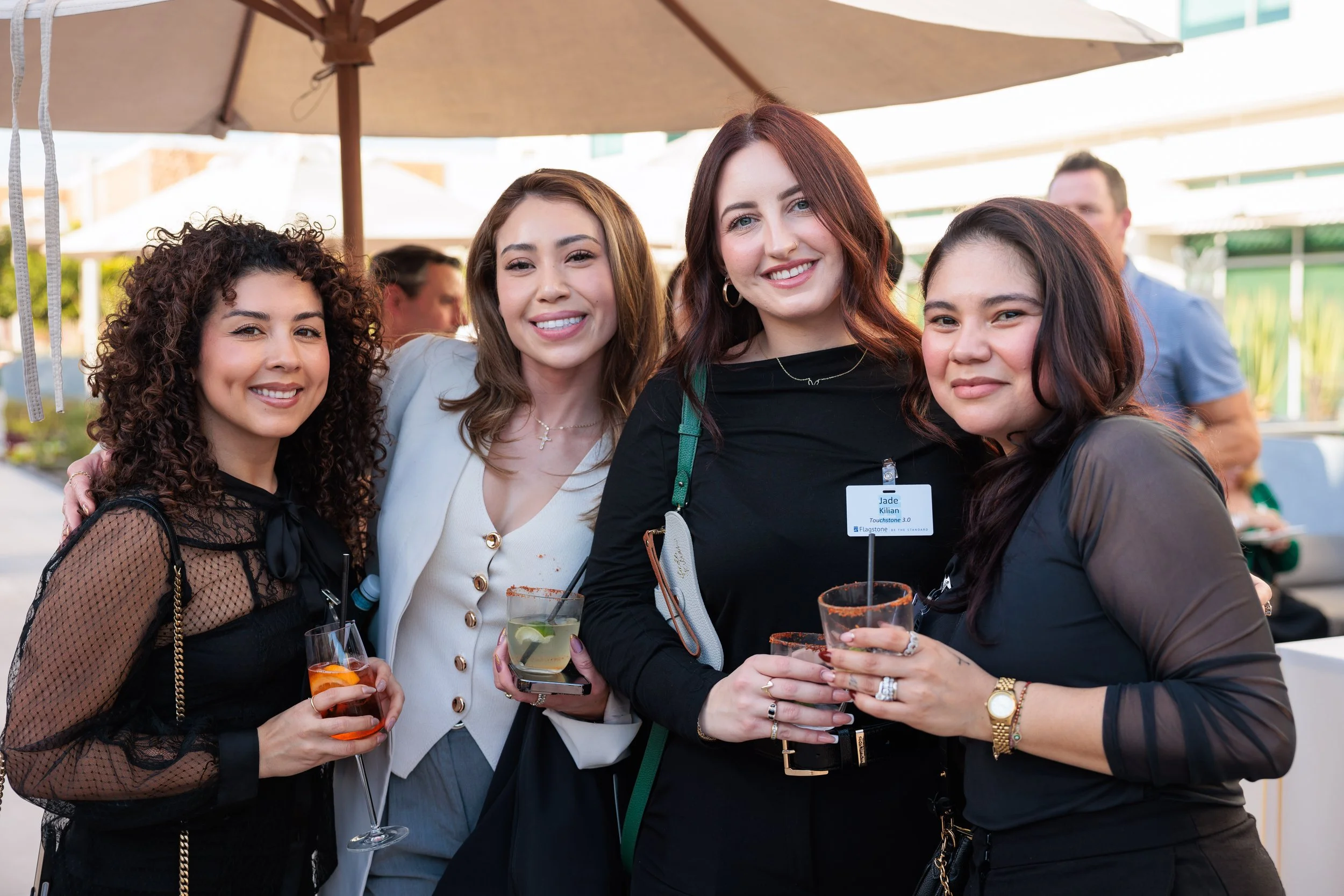 Four women at a social event holding drinks, smiling at the camera, with a large umbrella and other people in the background.