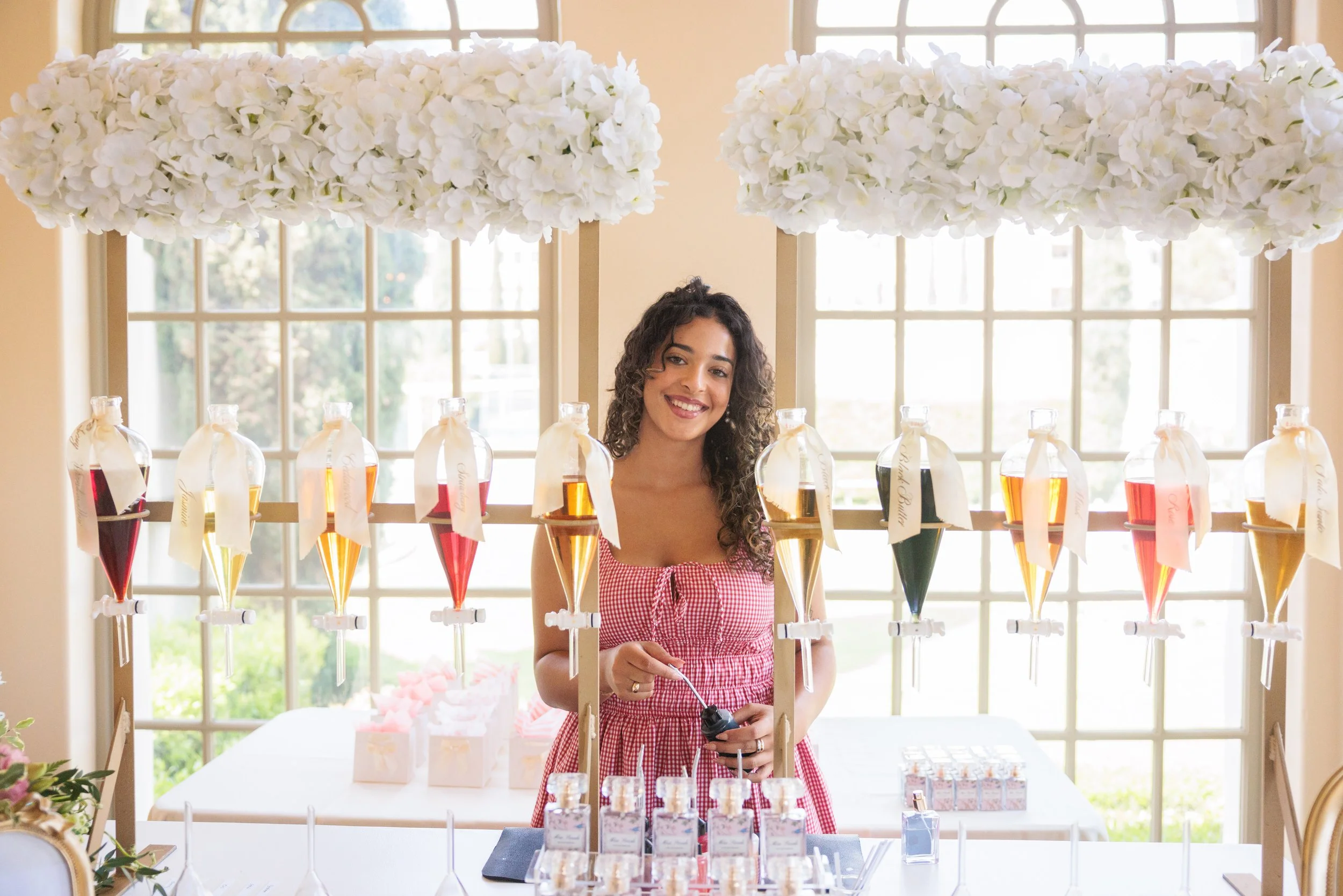 Young woman at a colorful syrup stand with multiple glass bottles containing different flavored syrups, smiling, in a bright room with large windows and floral decorations.