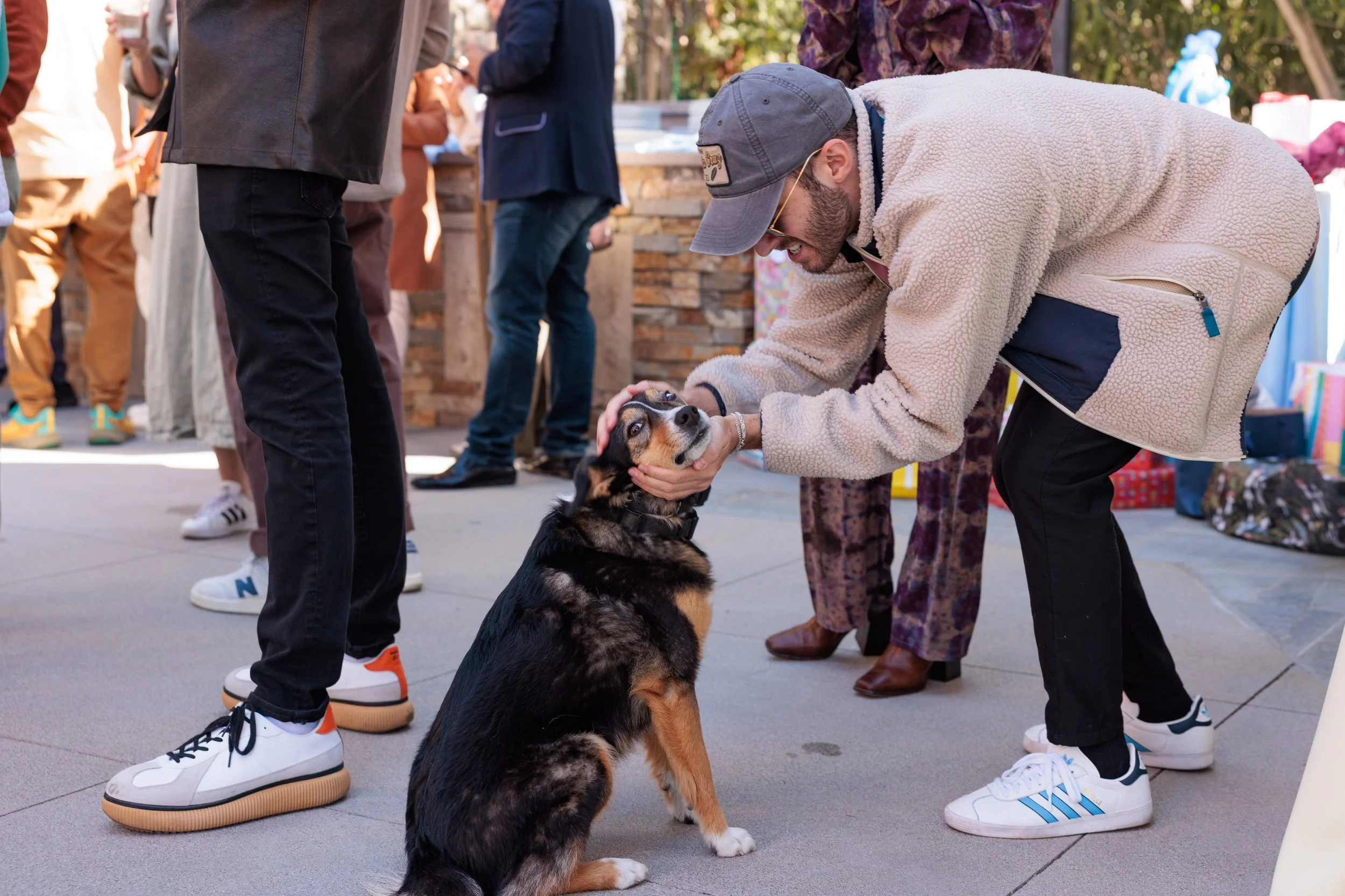 A man wearing a gray hat and beige fleece jacket gently holding a dog’s face while kneeling on the ground. The dog, a black and brown shepherd mix, is sitting and looking up at the man. Several people are standing around in an outdoor setting with so