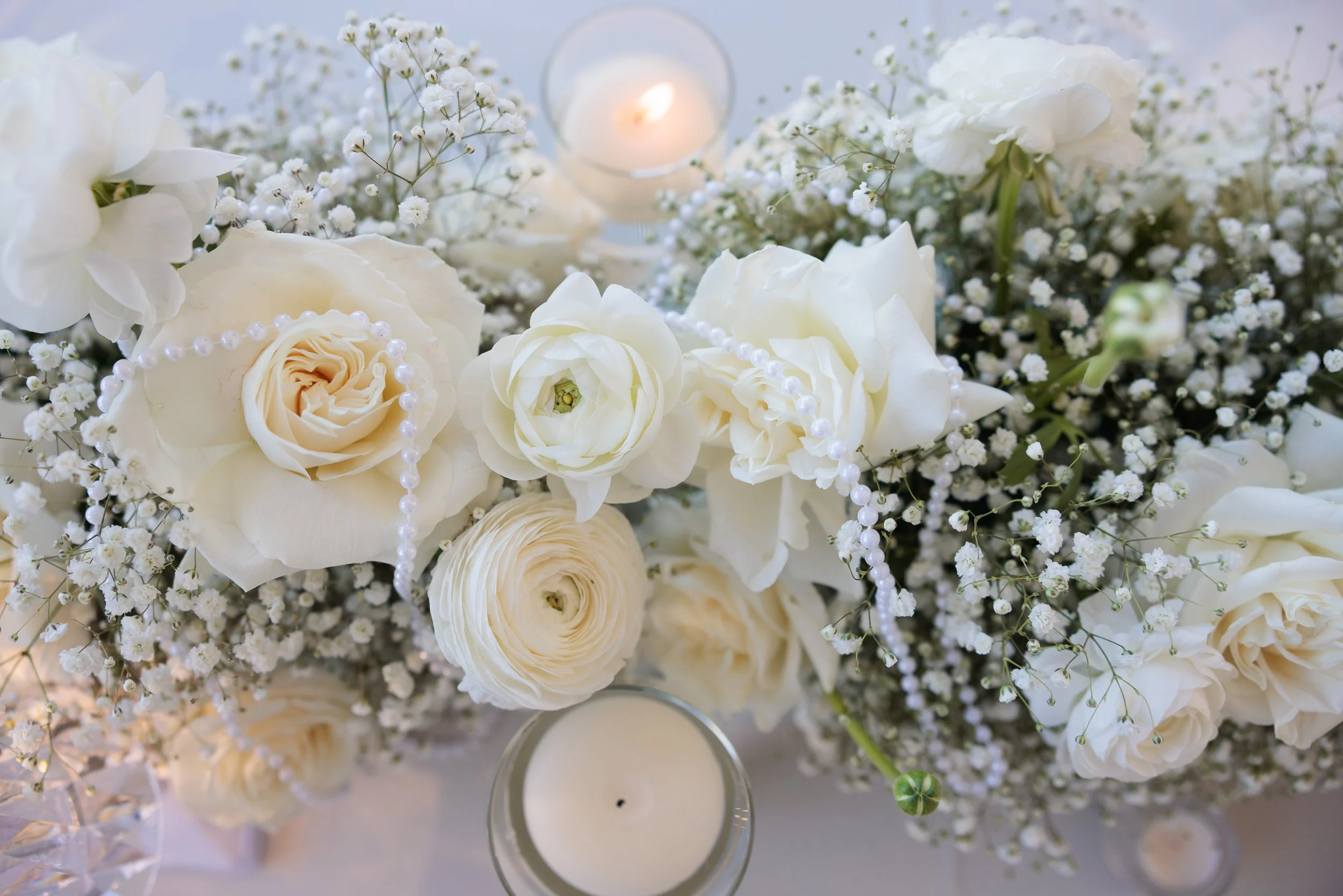 A floral arrangement with white roses, ranunculus, baby's breath, and beads, with a lit candle in the background.