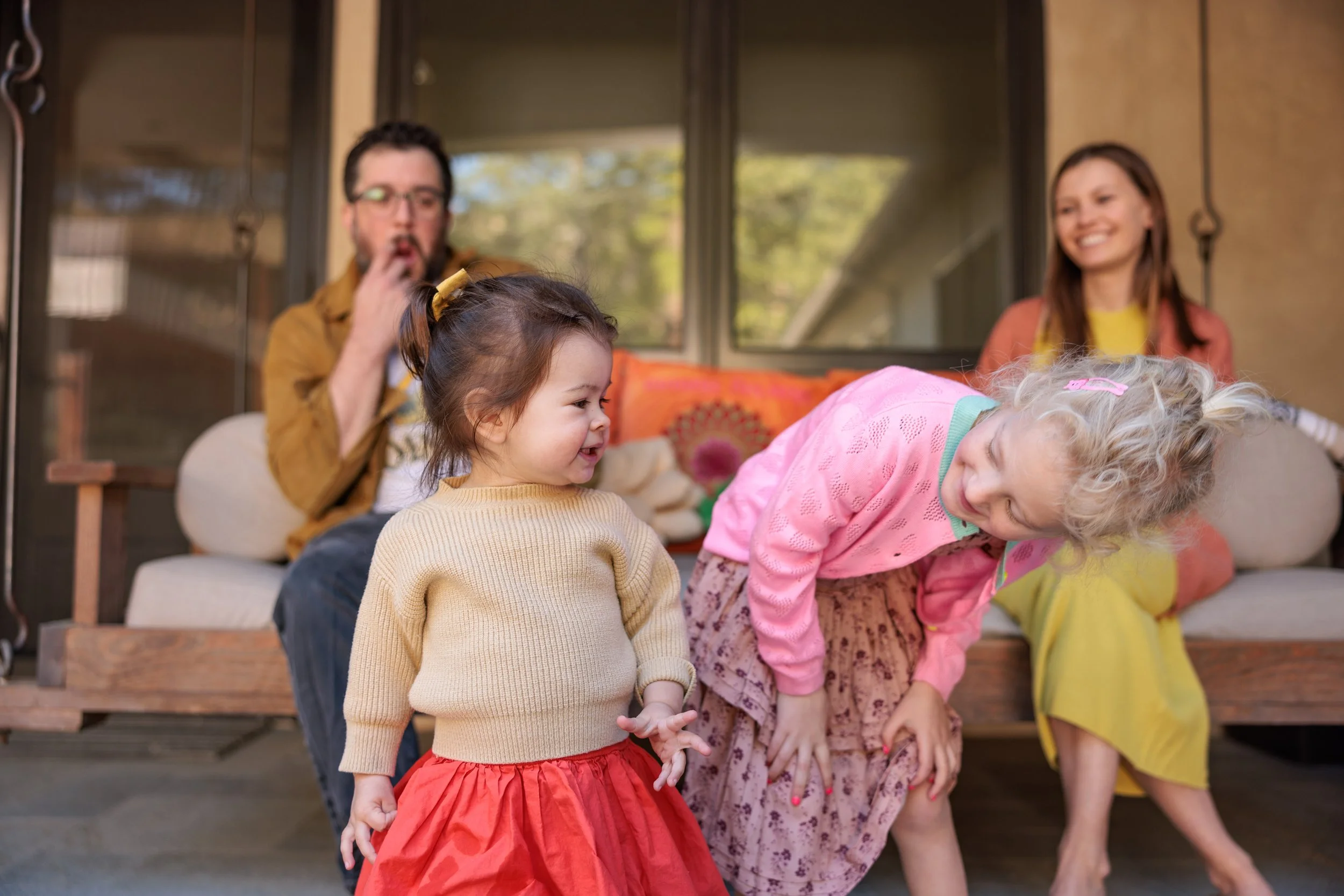 Four people, two young girls and two adults, sitting on a porch. The girls are laughing and playing, with one girl in a beige sweater and red skirt and the other in a pink sweater and floral skirt. The adults, a man with glasses in a brown jacket and