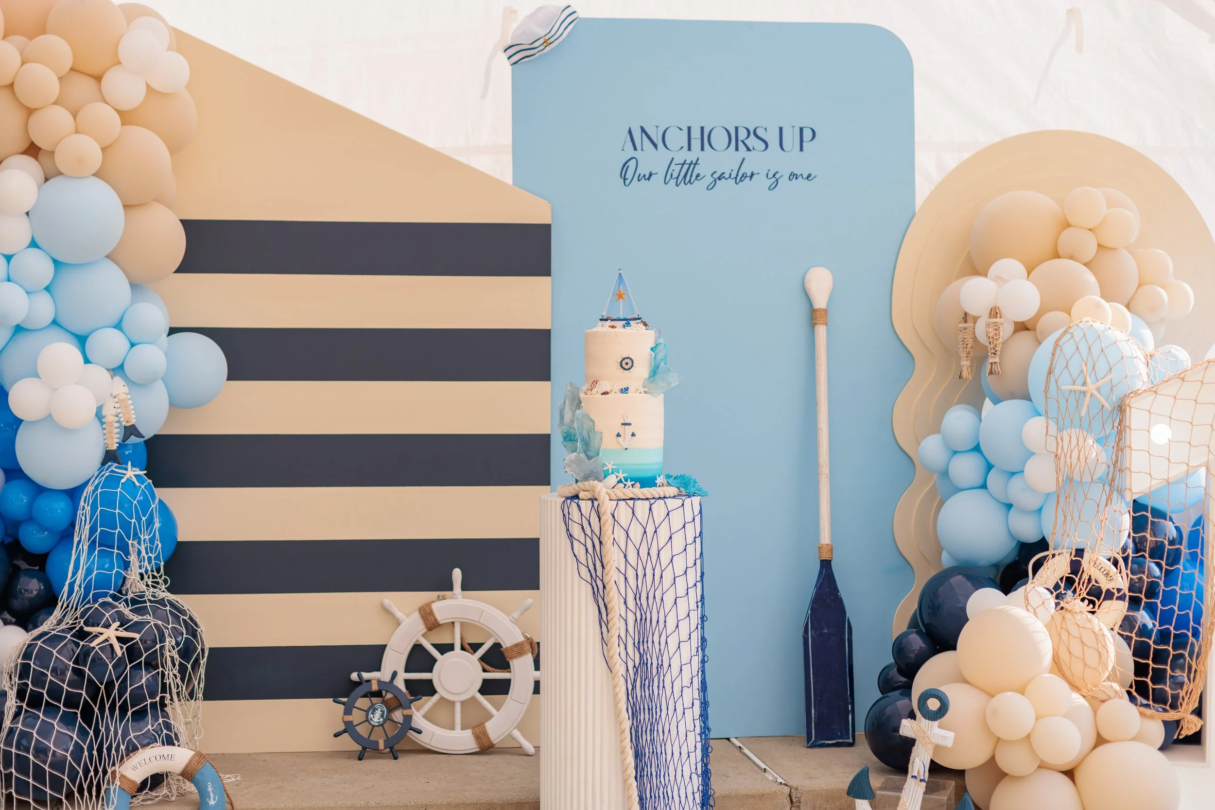 Decorative nautical themed backdrop with a striped blue and beige wall, balloon arrangements in blue, white, and beige, a cake with ocean and sailor motifs, a large blue and beige paddle, a ship's wheel, and a sign with the text "Anchors Up, Our litt