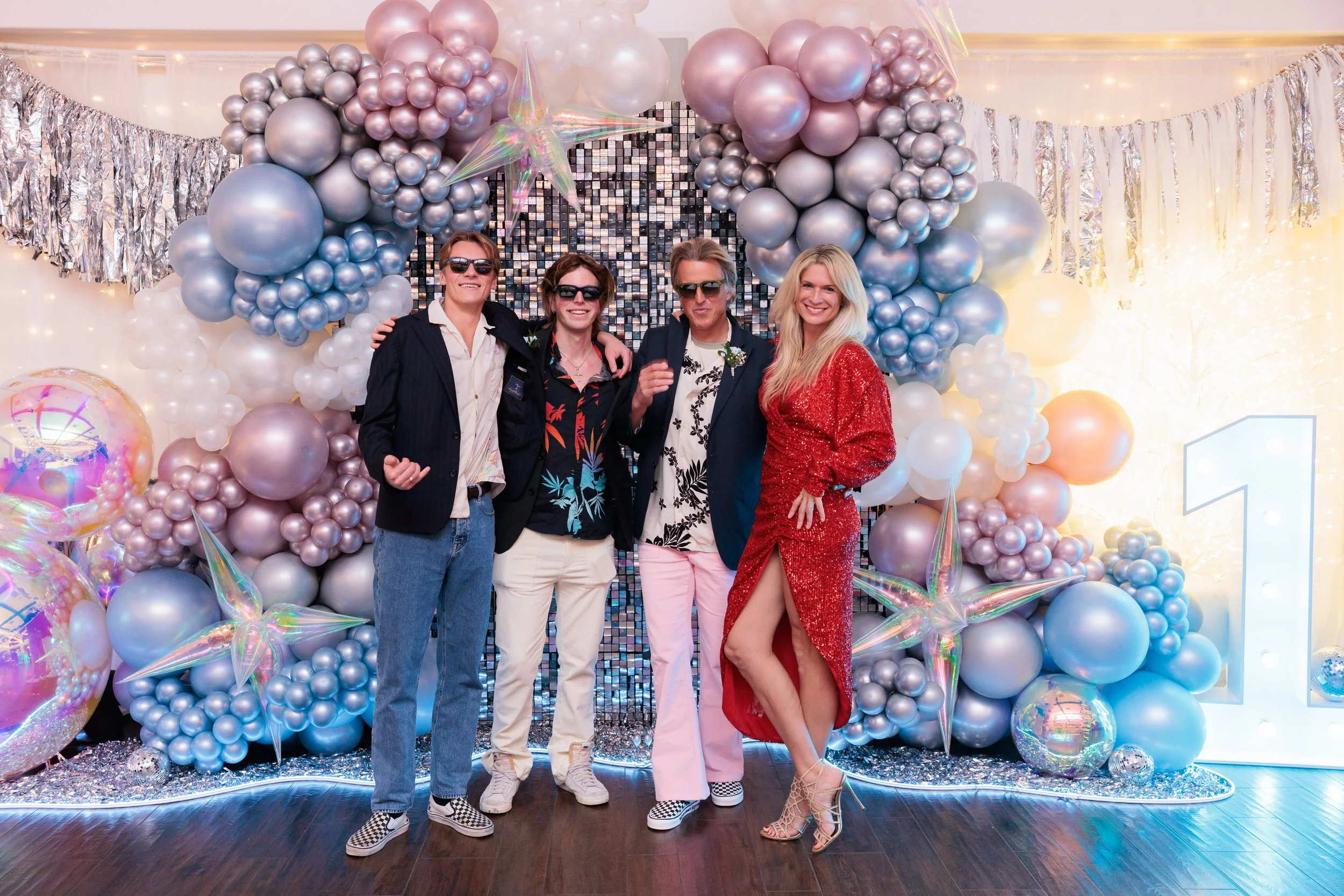 Group of four people dressed in party attire, standing in front of decorative balloon backdrop with silver, pink, and blue balloons, shiny star-shaped balloons, and a large illuminated number one, celebrating a special occasion.