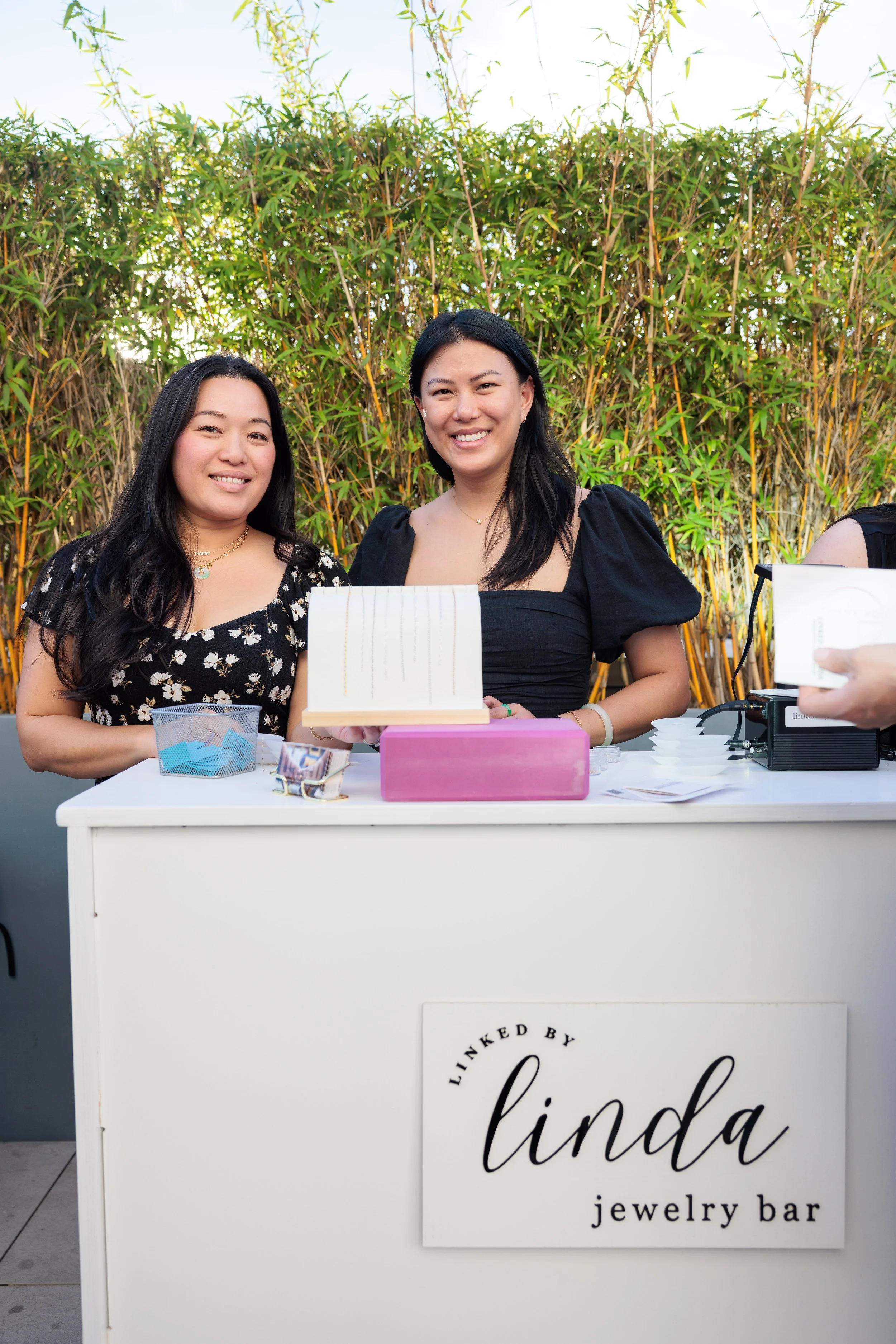 Two women smiling at a jewelry bar with a sign that says 'Linked by Linda jewelry bar,' outdoors with lush green bamboo behind them.