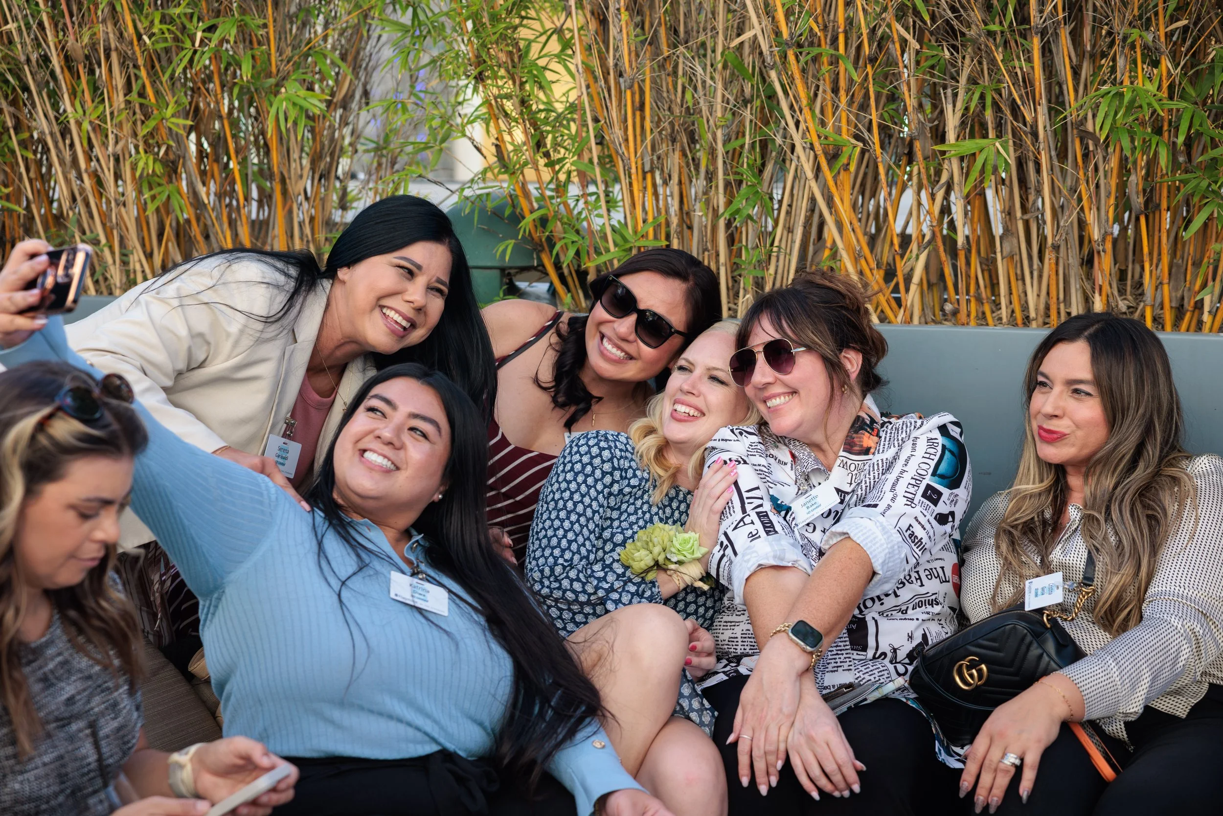 Group of women taking a selfie together outdoors, smiling and wearing sunglasses, with bamboo plants in the background.