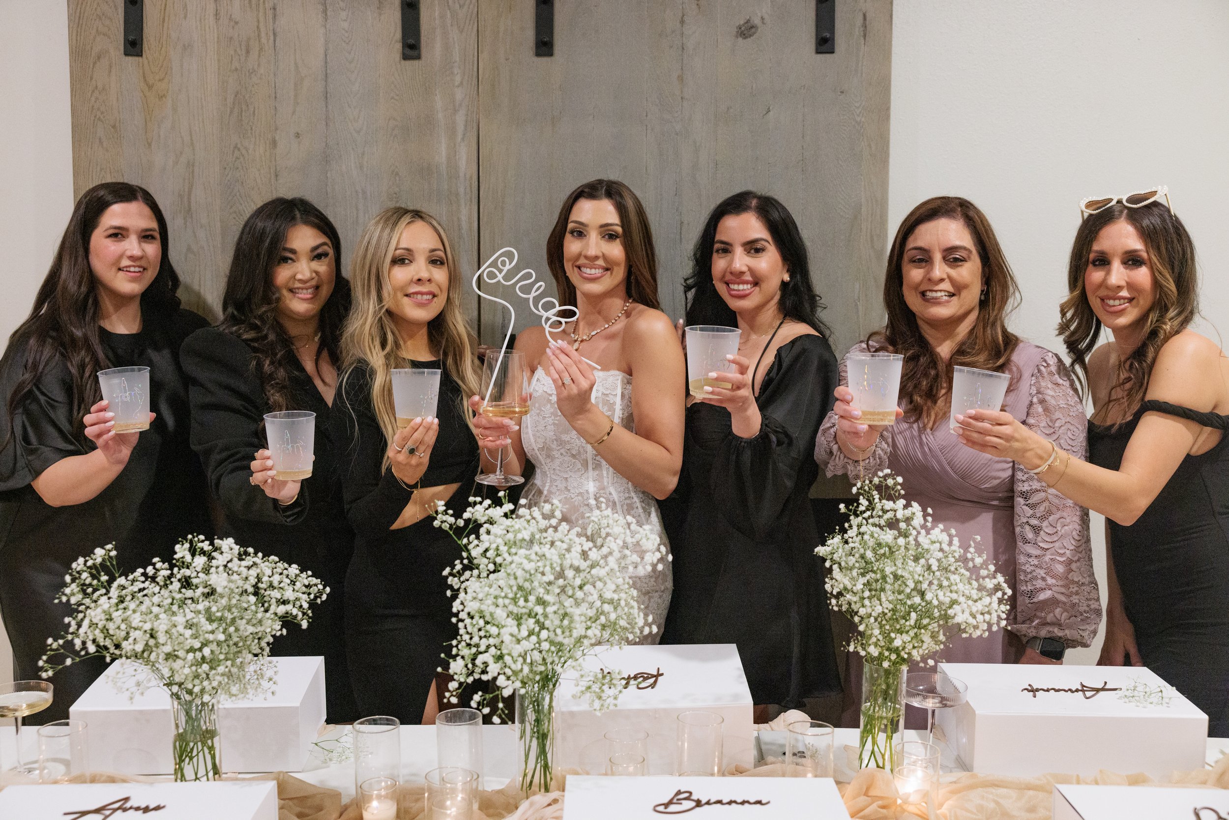 Group of eight women at a celebration, standing behind a table with white flowers and gift boxes, holding drinks, smiling at the camera.
