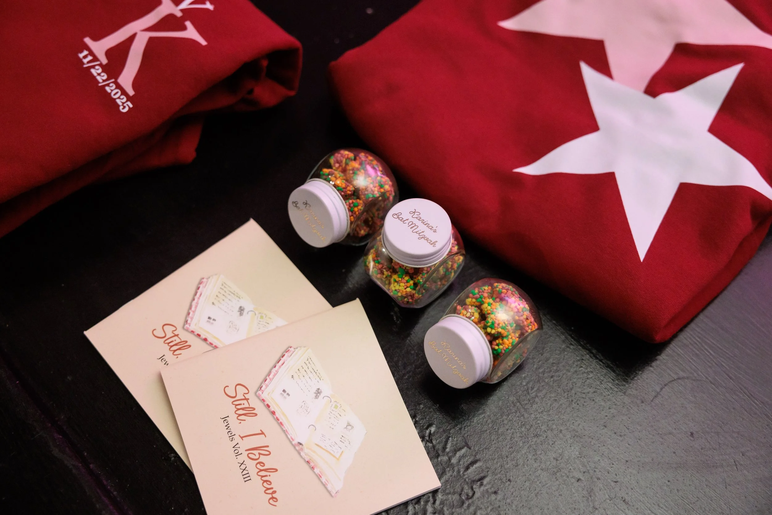 Two Canadian flags in folded fabric, three jars of multicolored sprinkles, and two wedding programs titled "Still I Believe" placed on a dark surface.