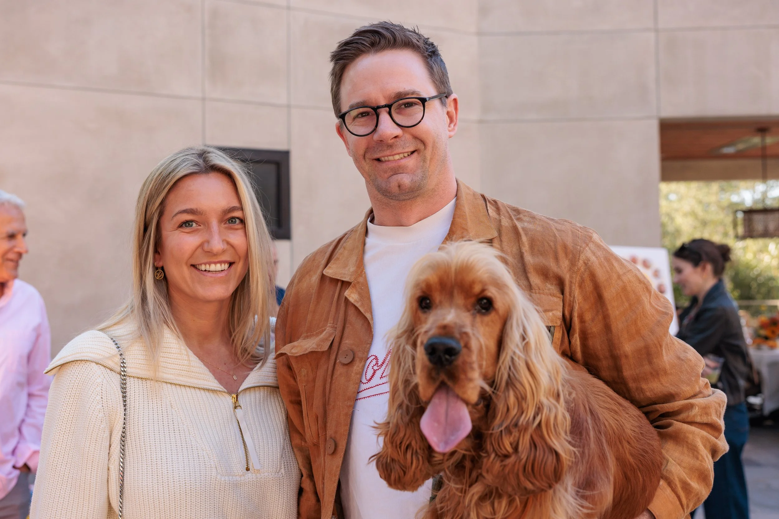 A smiling couple, a man with glasses and a woman with blonde hair, standing outdoors with a golden retriever dog in the man's arms, at a social gathering or event.