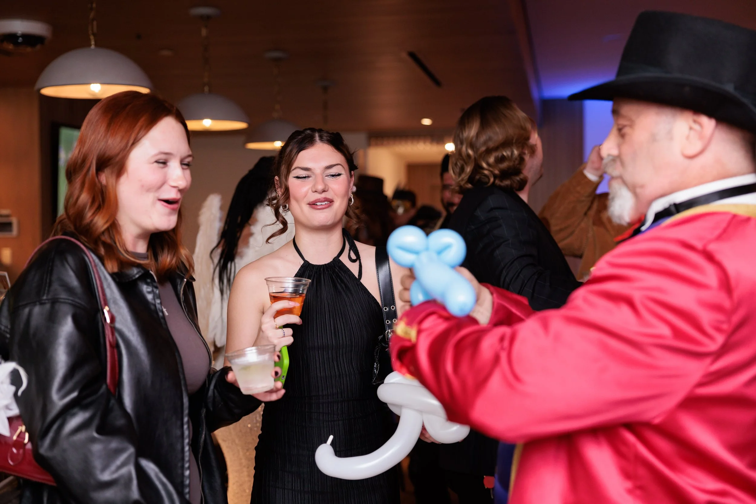 People at an event talking to a performer dressed as a clown who is holding a balloon animal.