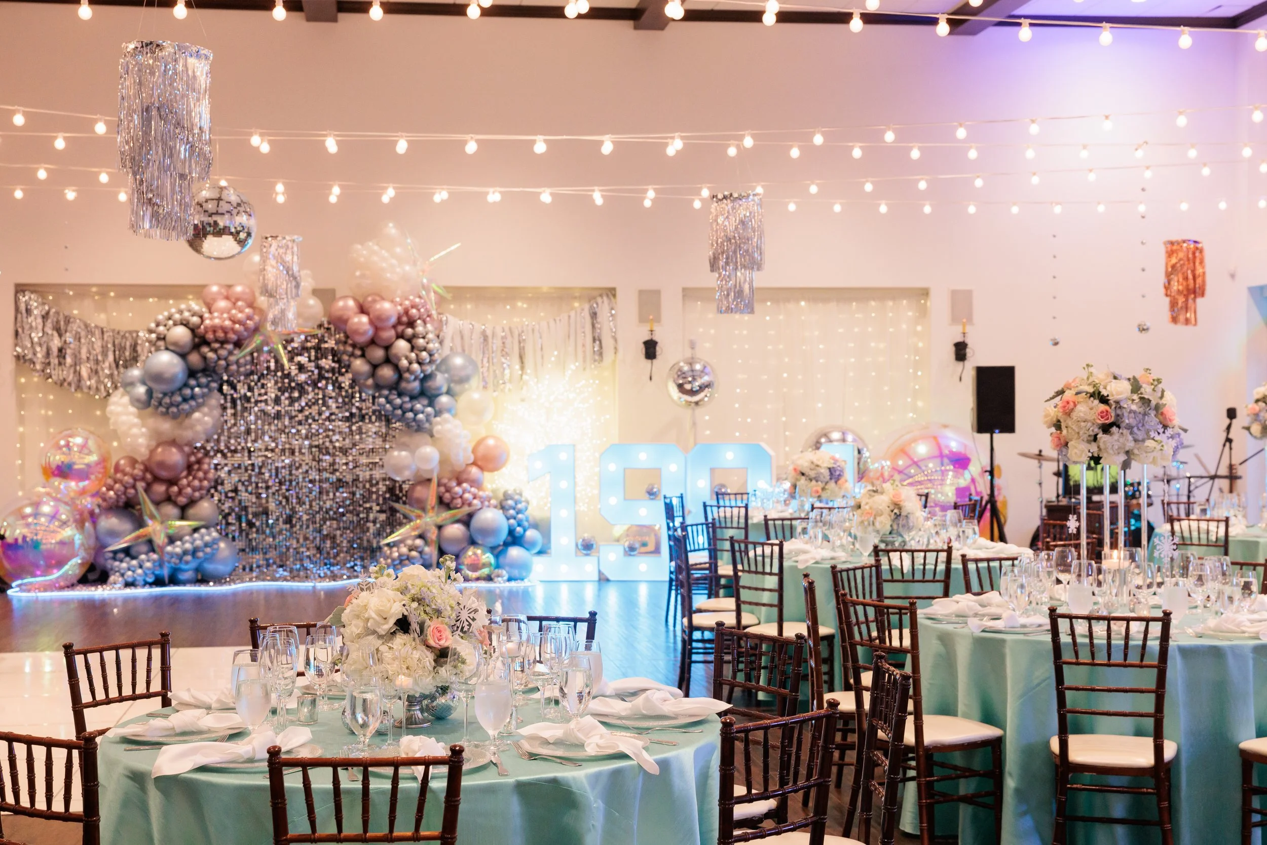 Celebration event decorated with balloons, fairy lights, and floral arrangements, featuring a lit-up '100' sign on a decorated stage with tables set for dining.