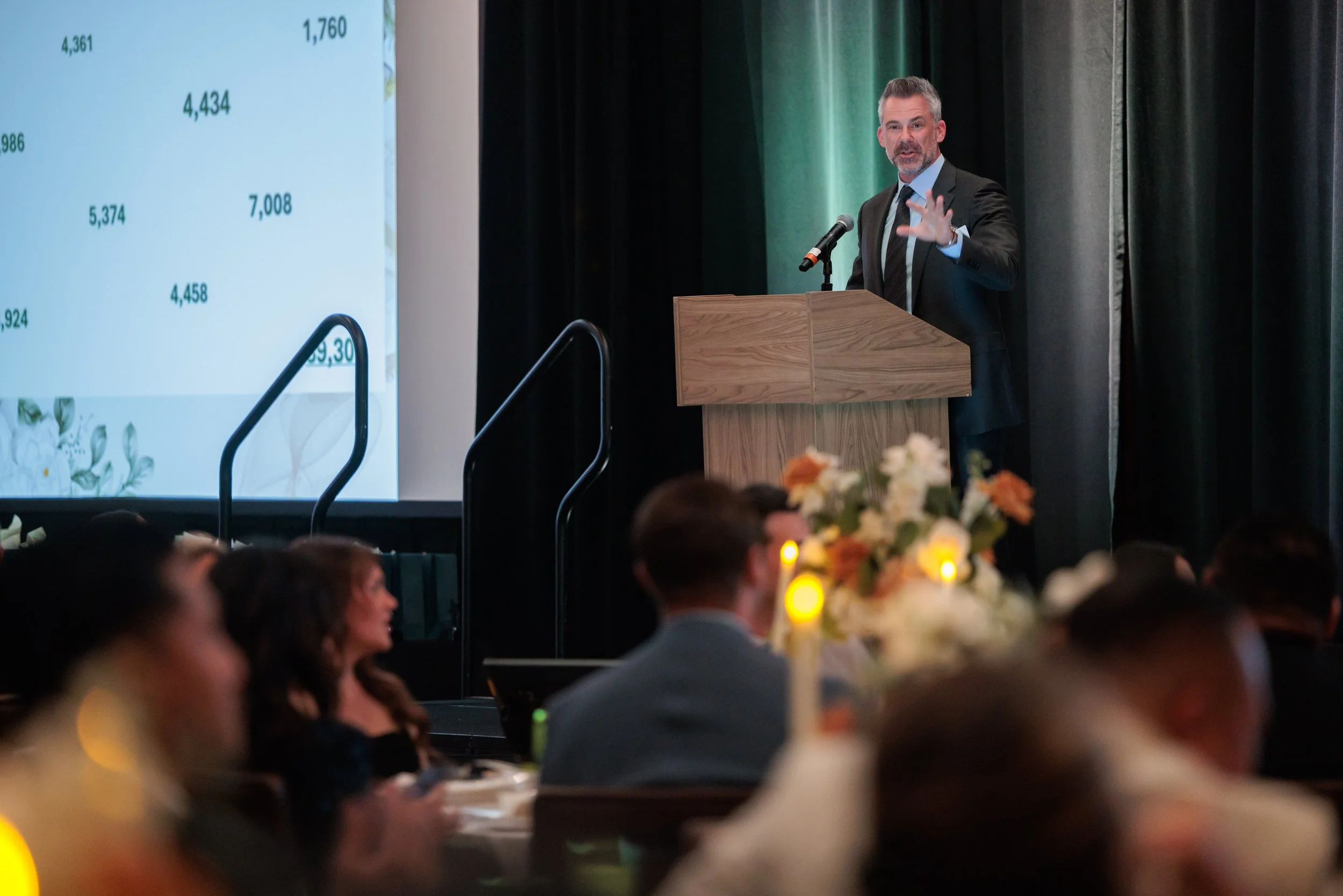 Man in suit giving a presentation at a podium on stage, with a large screen displaying data behind him, audience seated at tables with floral centerpieces and candles.