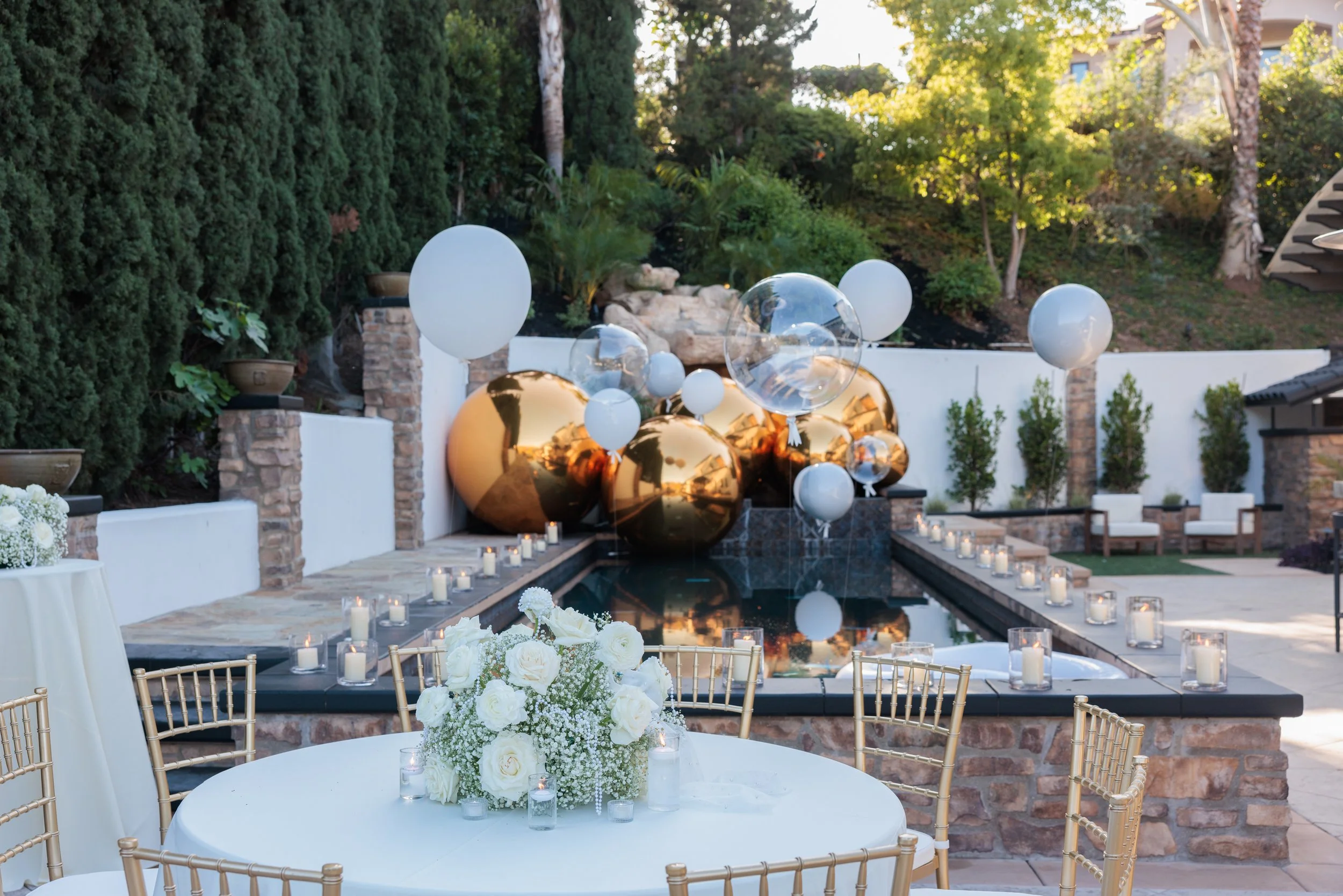 Decorative outdoor event setup featuring a round table with white flowers and candles, a pool with large gold and transparent balloons, surrounded by chairs, candle lanterns, and lush greenery.