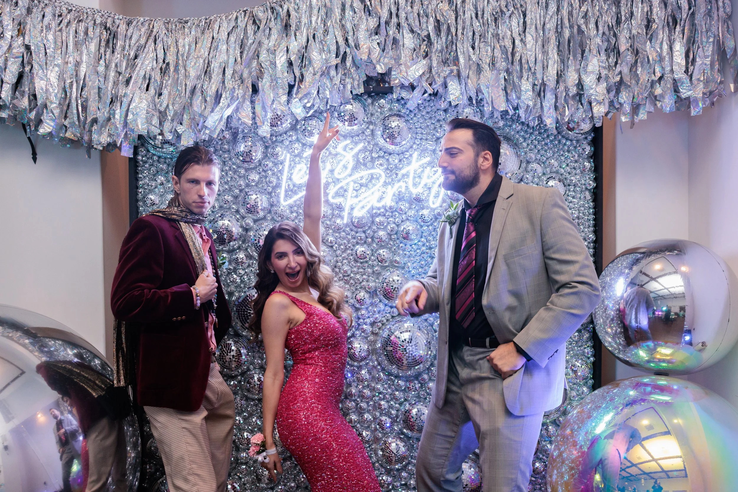 Four people posing at a party with a shiny, silver, disco-themed backdrop that has the words 'Let's Party' illuminated in neon. The group includes two men in suits and a woman in a pink, glittery dress, with large reflective balloons around them.