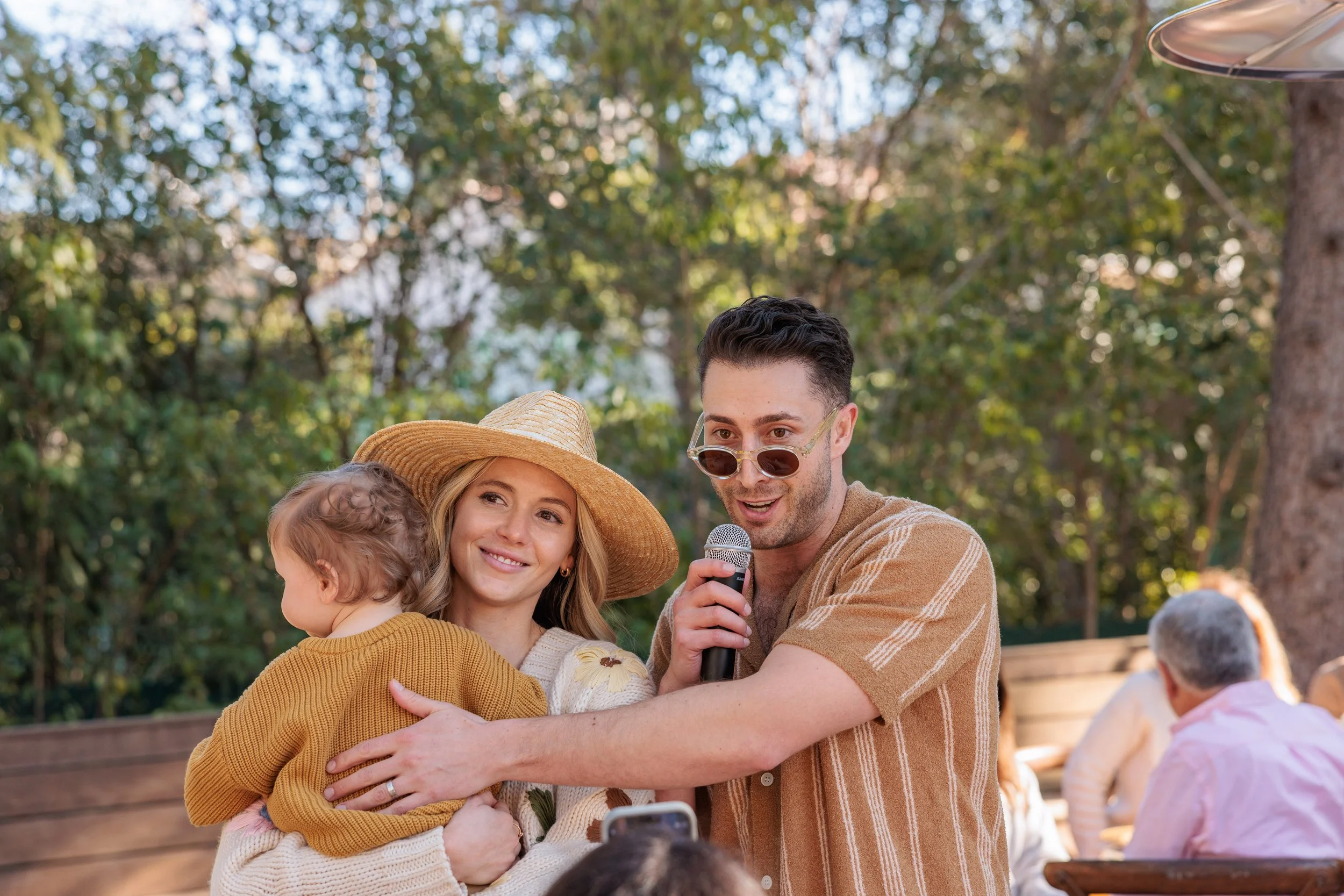 A young man wearing sunglasses and a striped tan shirt holding a microphone is hugging a woman holding a toddler. The woman is wearing a wide-brimmed straw hat and a cream-colored sweater. They are outdoors with trees in the background and other peop