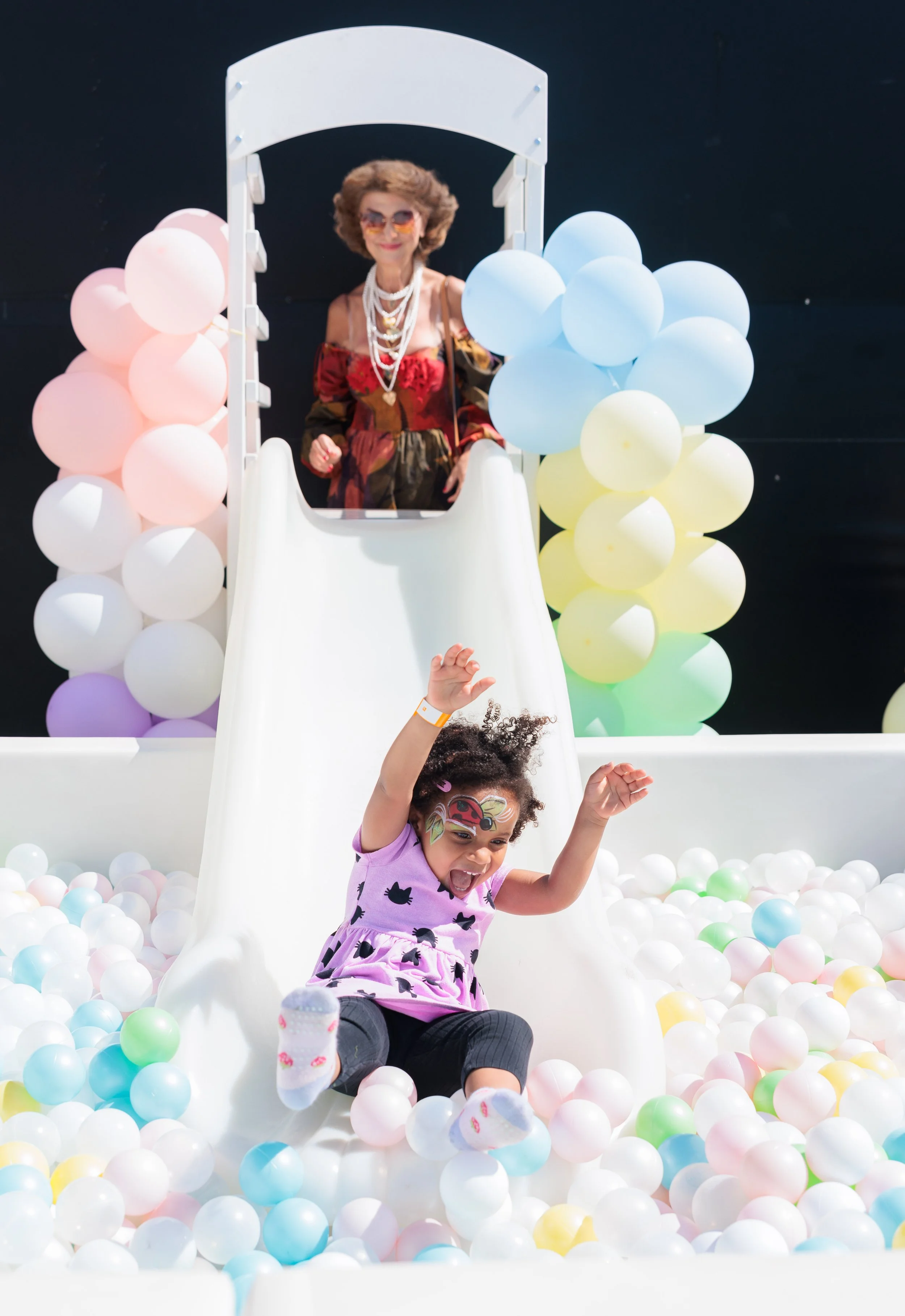 A young girl with face paint sliding down a white slide into a ball pit filled with colorful plastic balls, with an older woman standing at the top of the slide surrounded by balloons.