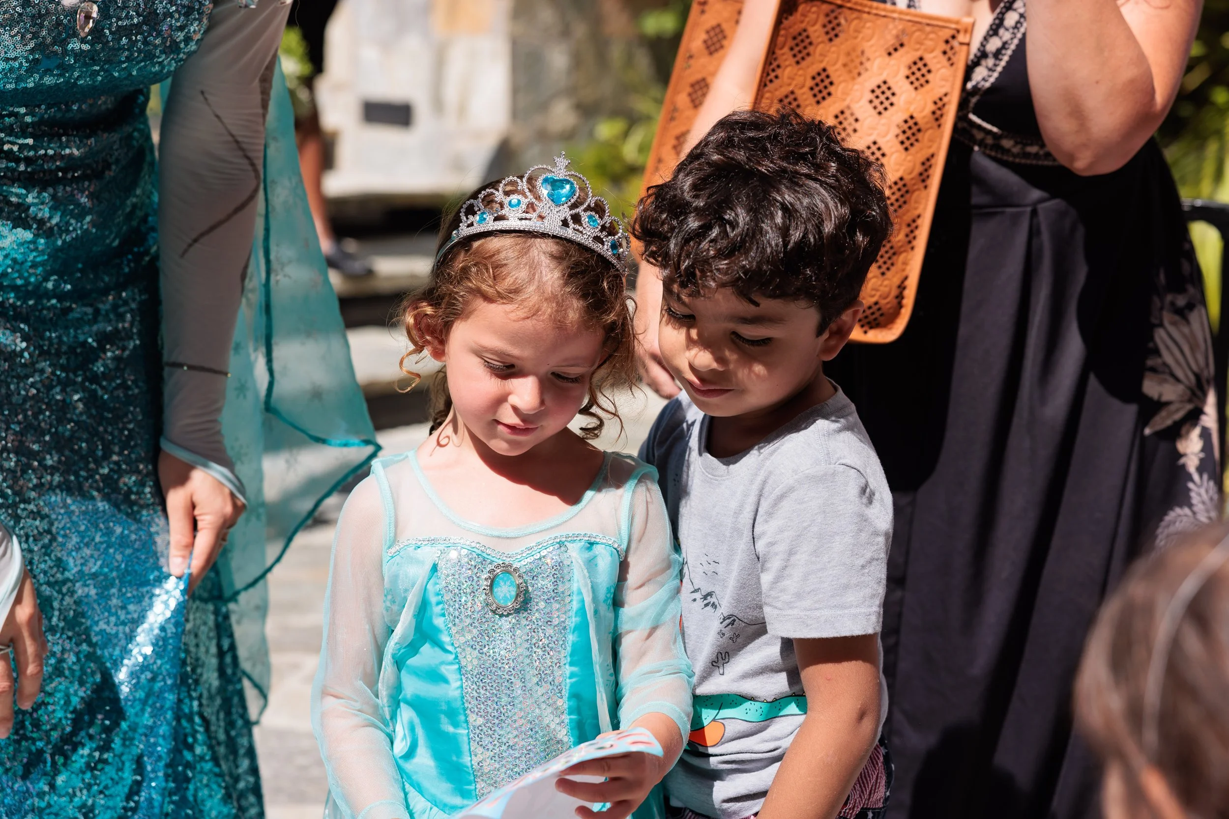 A young girl dressed as a princess and a young boy are looking at a piece of paper together. The girl is wearing a blue princess dress and a tiara, and the boy is wearing a gray t-shirt. There are women dressed in fancy outfits around them.