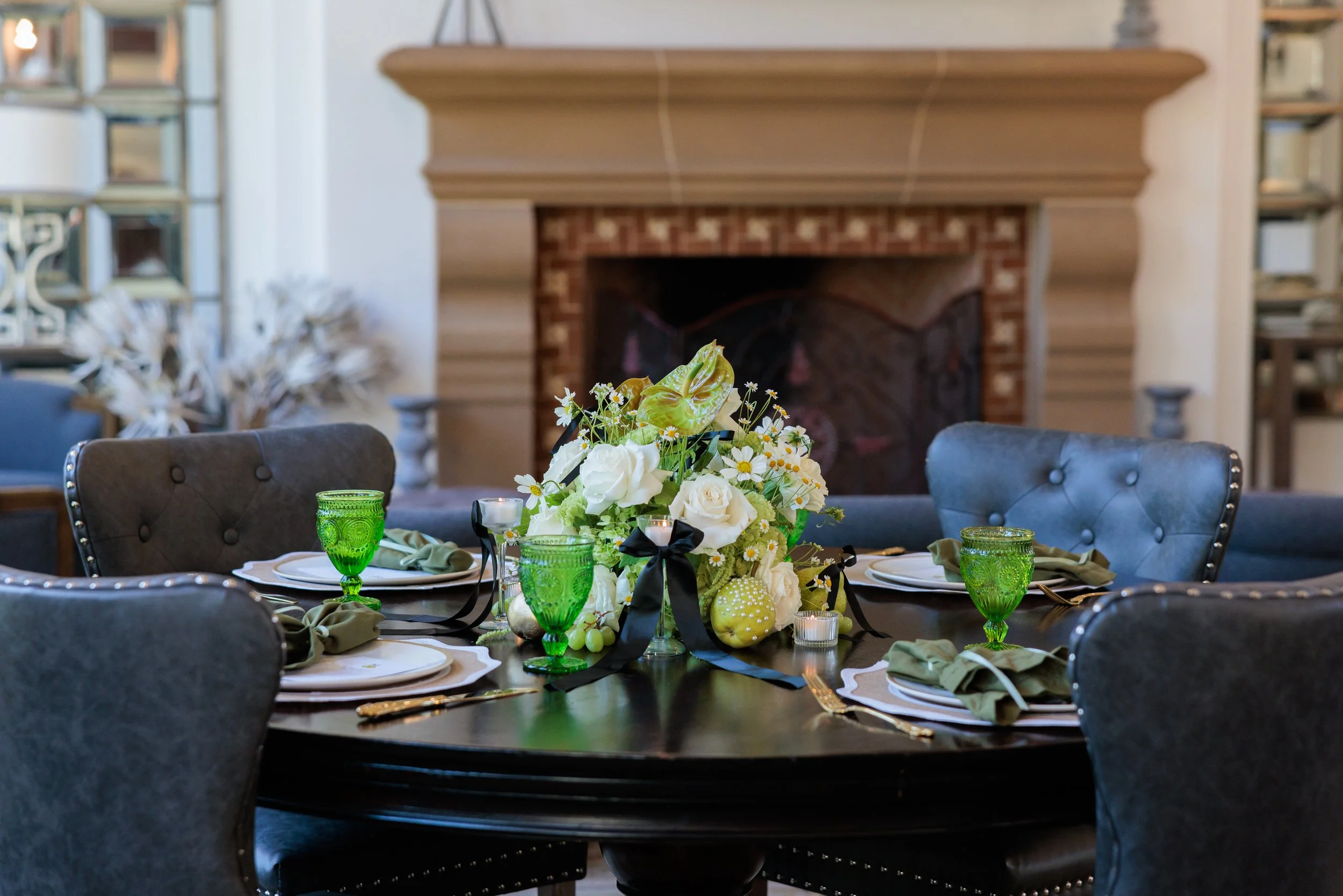 Round dining table set for a meal, with a floral centerpiece, green glasses, and colored napkins, in a room with a fireplace and shelves in the background.