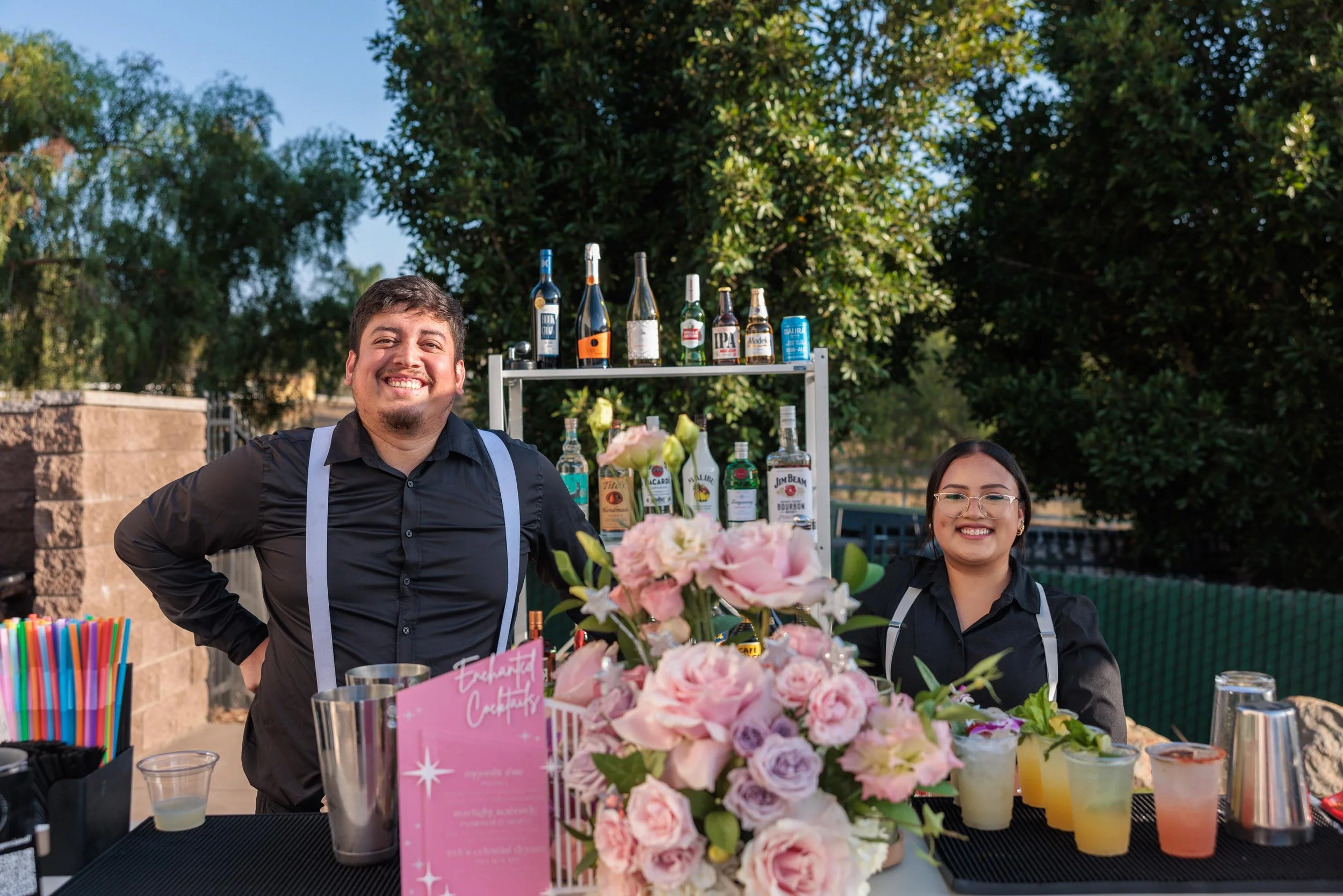 Two smiling bartenders standing behind a bar at an outdoor event with trees in the background. The male bartender has dark hair and a beard, wearing a black shirt with suspenders. The female bartender has dark hair, glasses, and is also wearing a bla