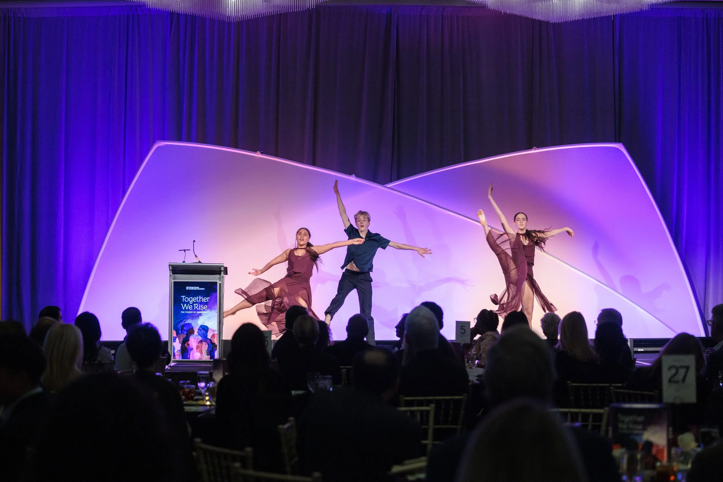 Three dancers performing on a stage with a colorful, curved backdrop, in front of an audience seated at tables.