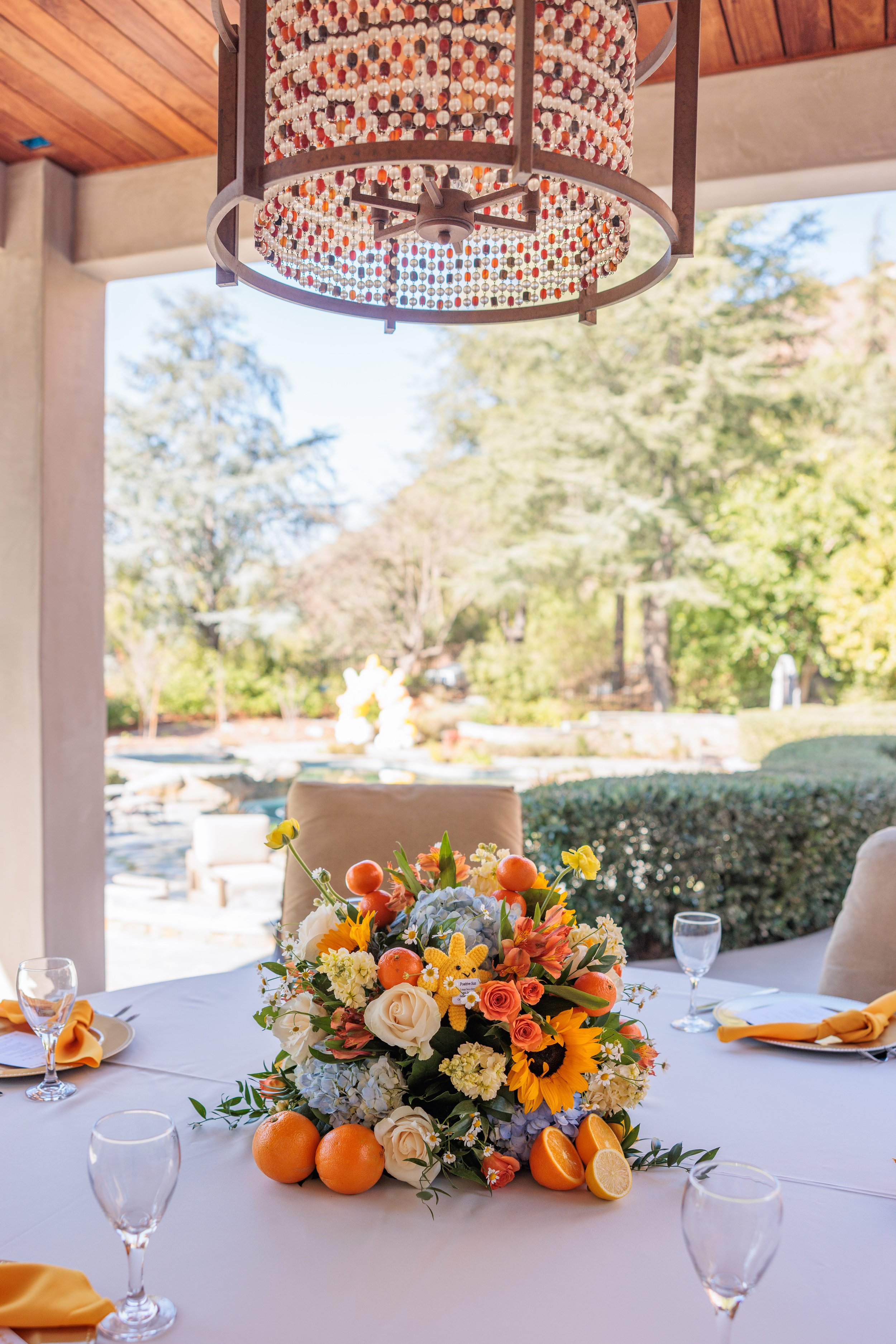 A decorated round table with a floral centerpiece of sunflowers, roses, hydrangeas, and other flowers, surrounded by oranges and lemon halves, with water glasses and yellow napkins, set in a bright room with large windows showing a garden or outdoor 