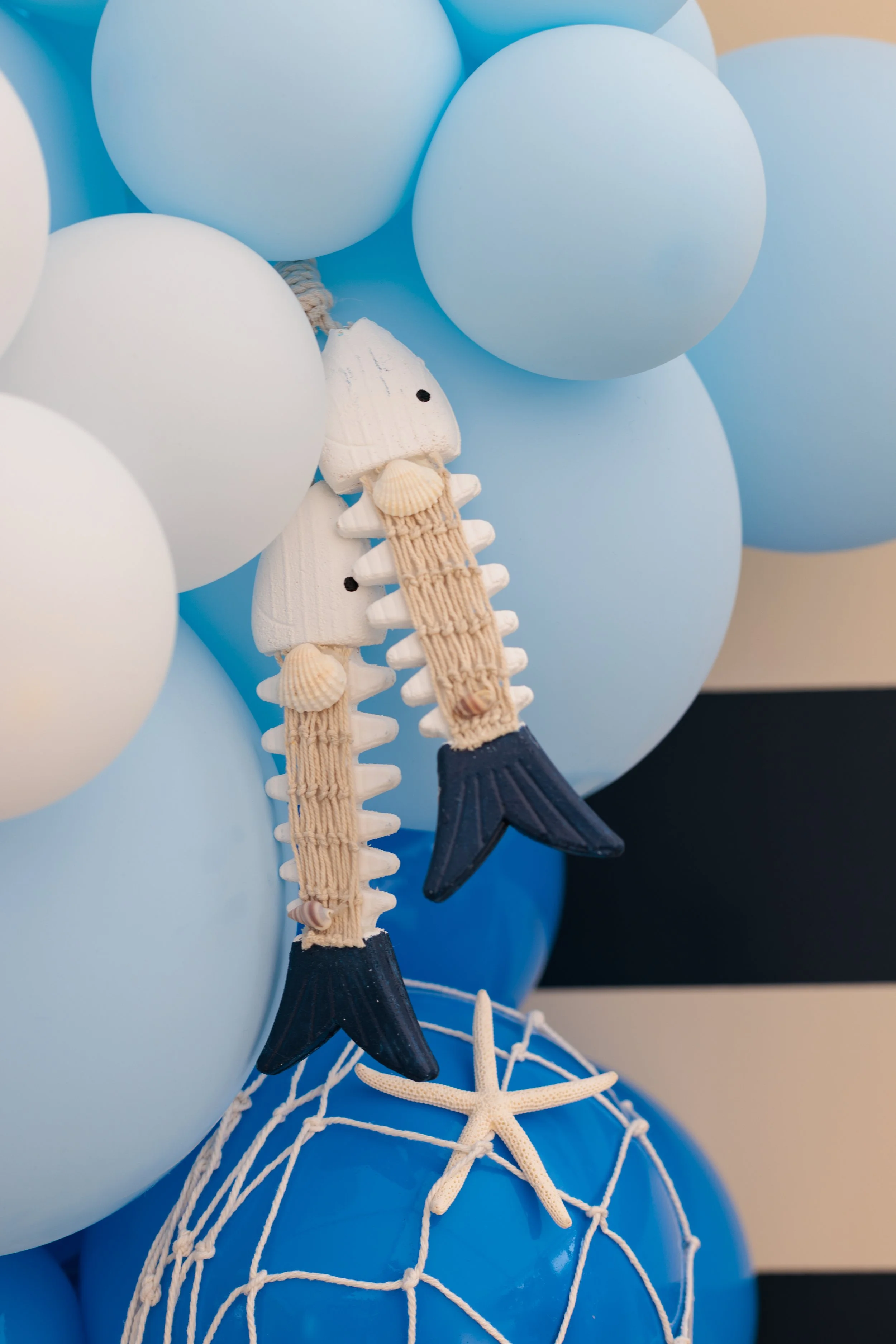 A cluster of light blue and white balloons with a decorative trinket resembling a mermaid tail and fish skeleton, accented with seashells and a small starfish.