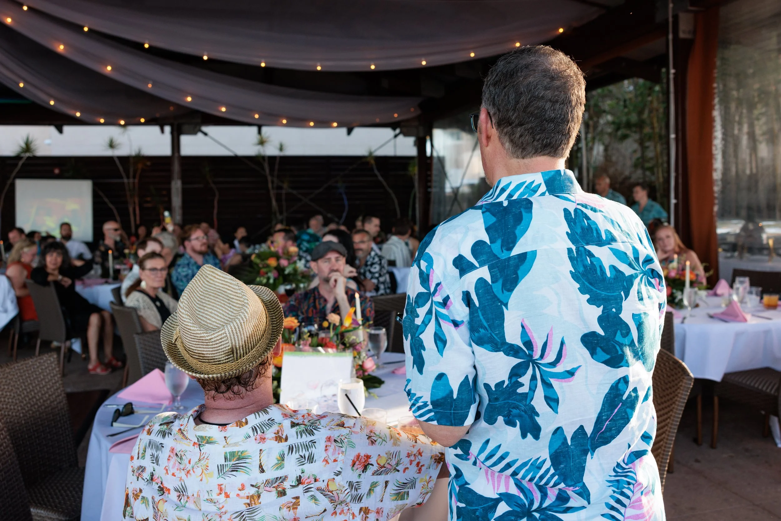 People at a social gathering or party, some wearing tropical shirts and hats, sitting at a decorated outdoor or semi-outdoor venue with tables, flowers, and a large crowd.