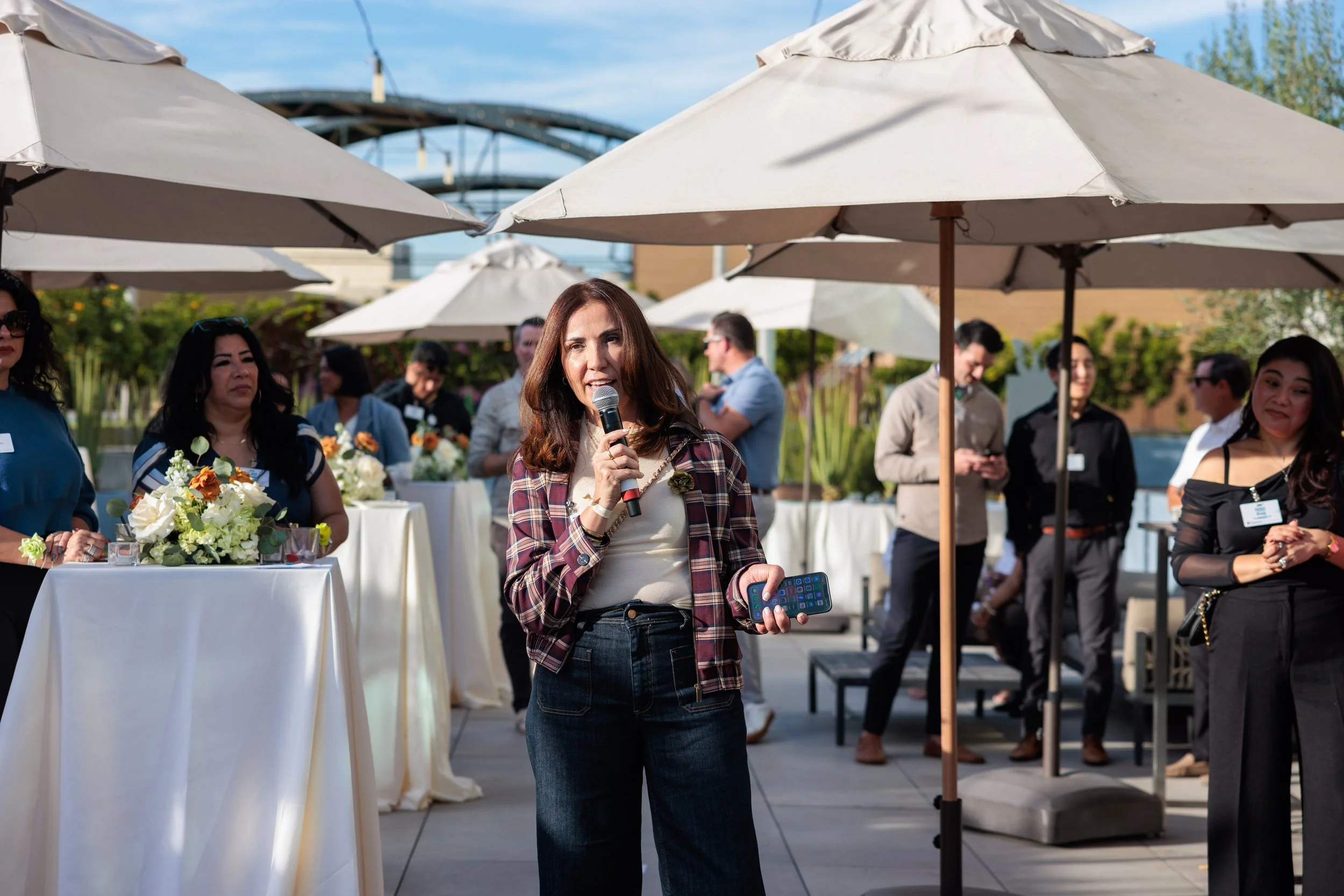 A woman with brown hair, wearing a plaid jacket, speaking into a microphone at an outdoor event. There are people around her under umbrellas, some with flowers and drinks on high tables.