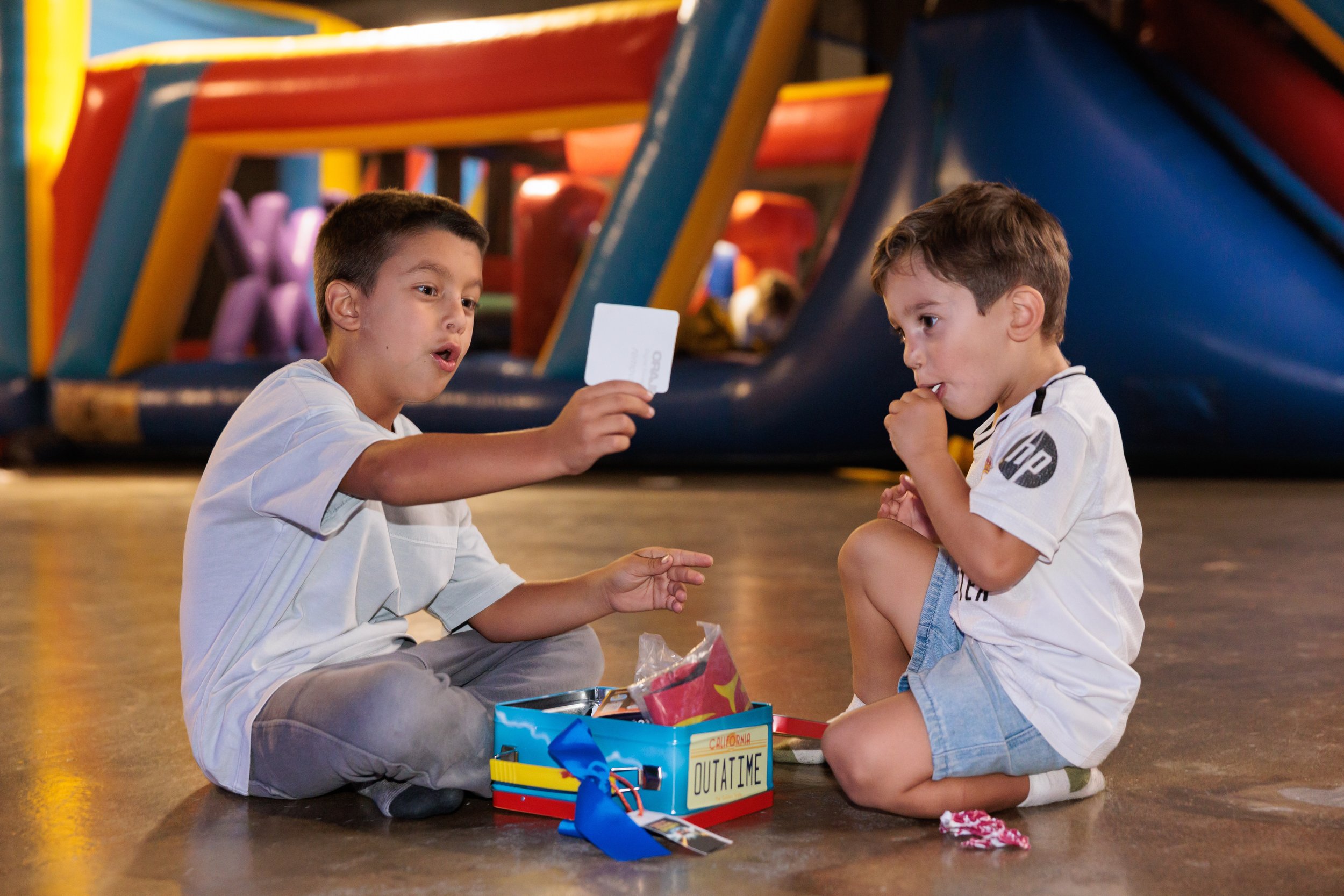 Two young boys sitting cross-legged on a wooden floor at an arcade or play center, playing a game with a card and tokens. One boy is holding a card and appears to be explaining something, while the other boy is watching with his thumb in his mouth. T