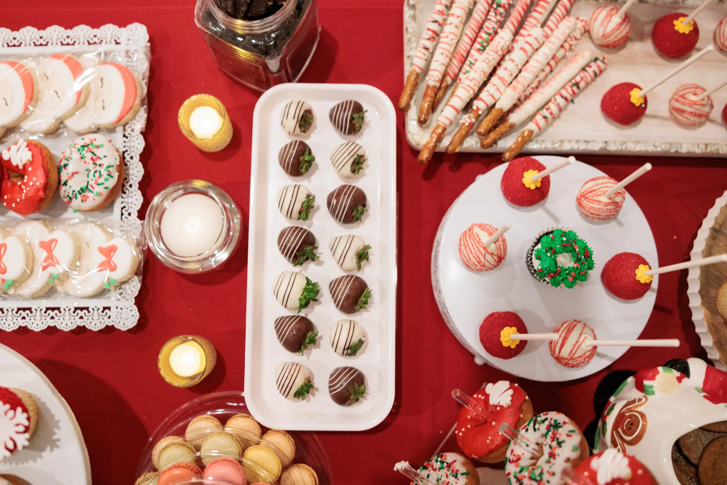 Festive holiday dessert table featuring decorated cookies, chocolate-covered strawberries, cake pops, and macarons on a red tablecloth with candles.