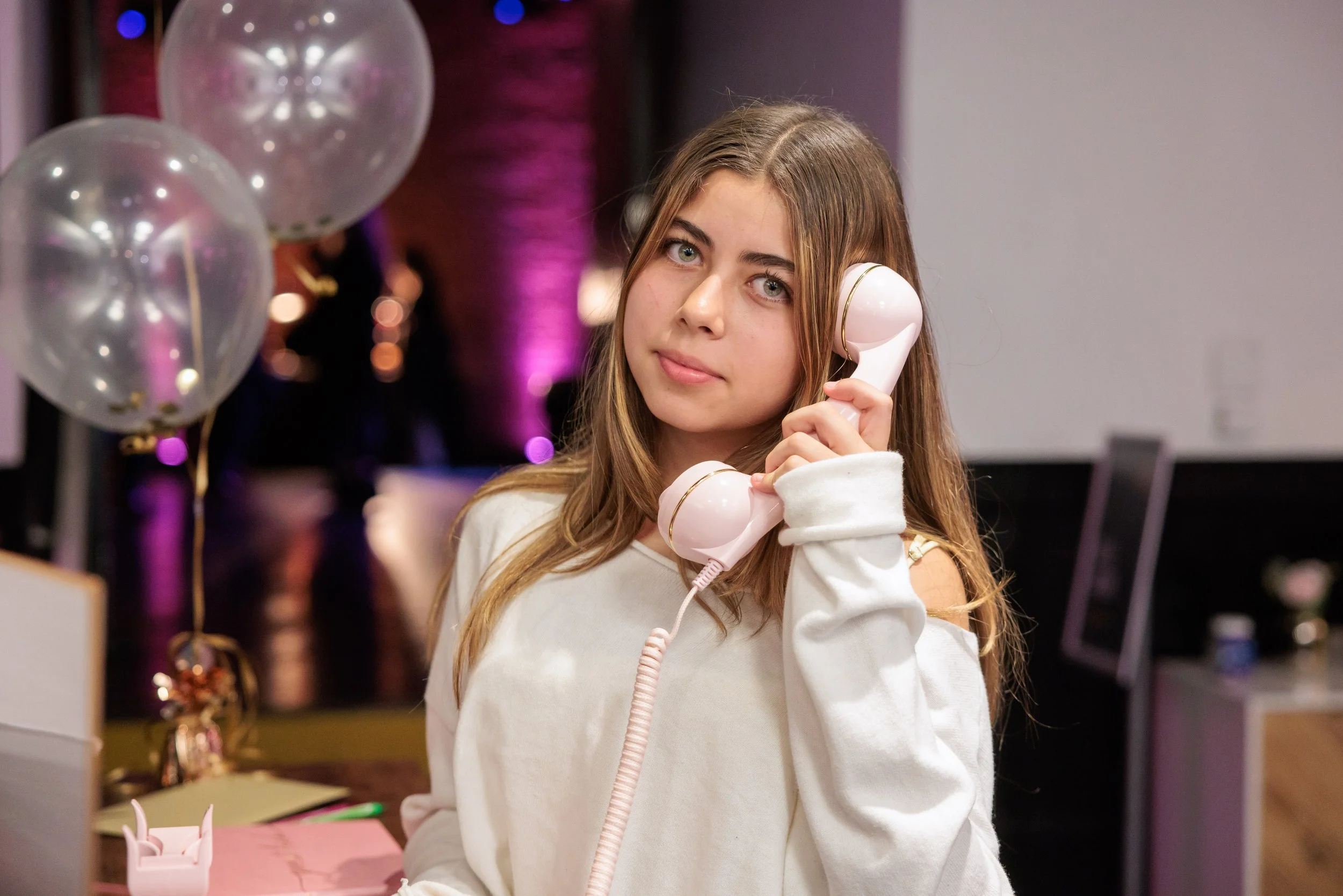 A young woman with long, light brown hair holding a vintage pink telephone receiver to her ear, standing indoors with pink and purple lighting, and balloons in the background.