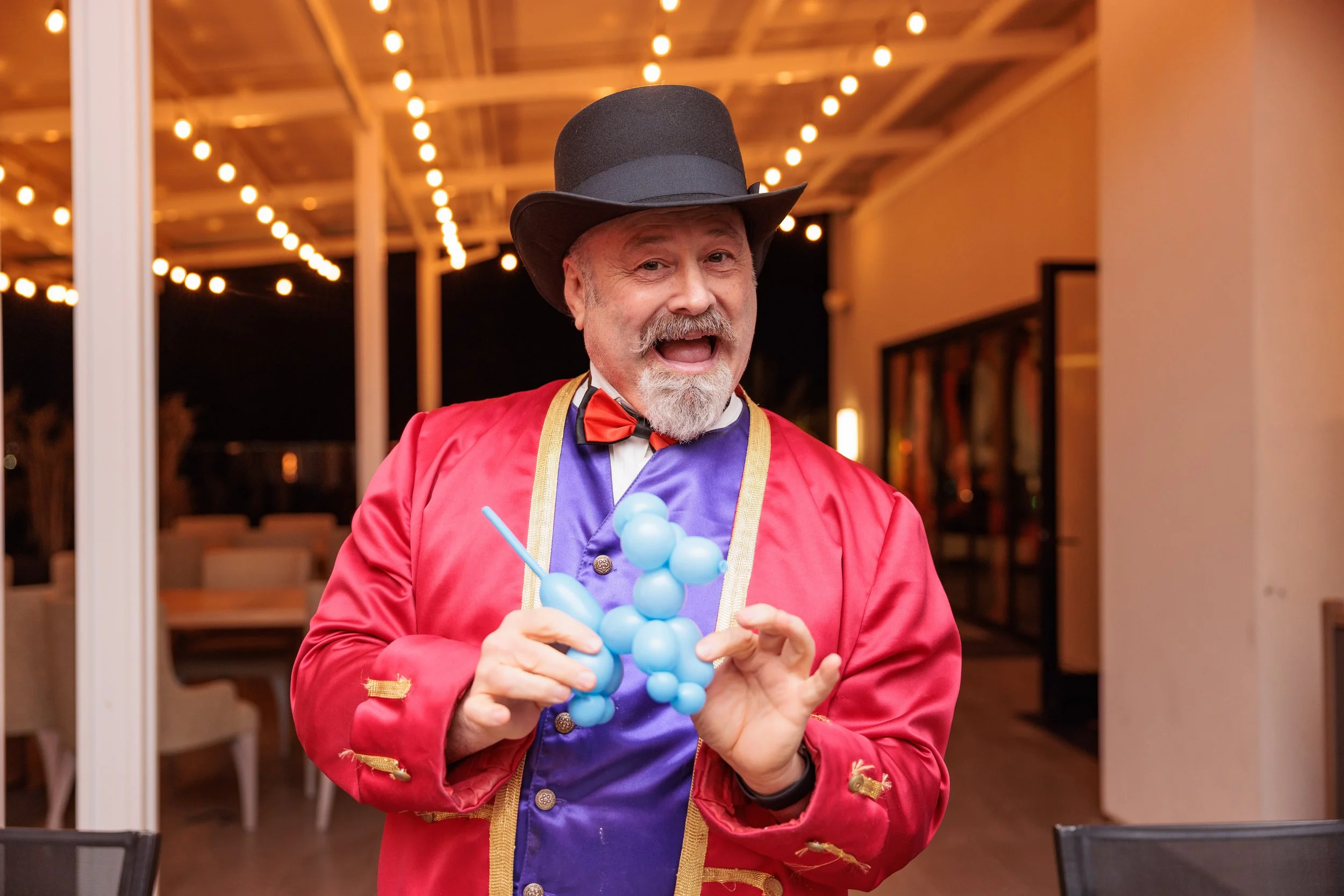 A man dressed as a circus or carnival performer with a black top hat, red coat, purple vest, and red bow tie, holding a blue balloon sculpture, standing in a decorated indoor space with string lights.