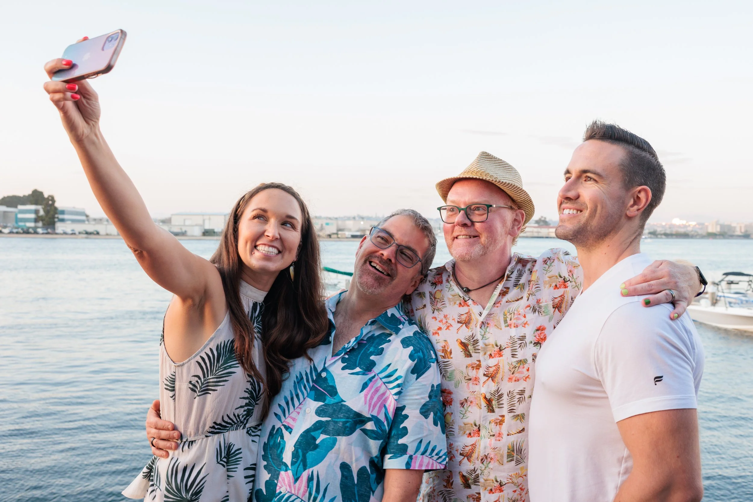 Four friends taking a selfie by the water on a clear day, smiling, with boats and buildings in the background.