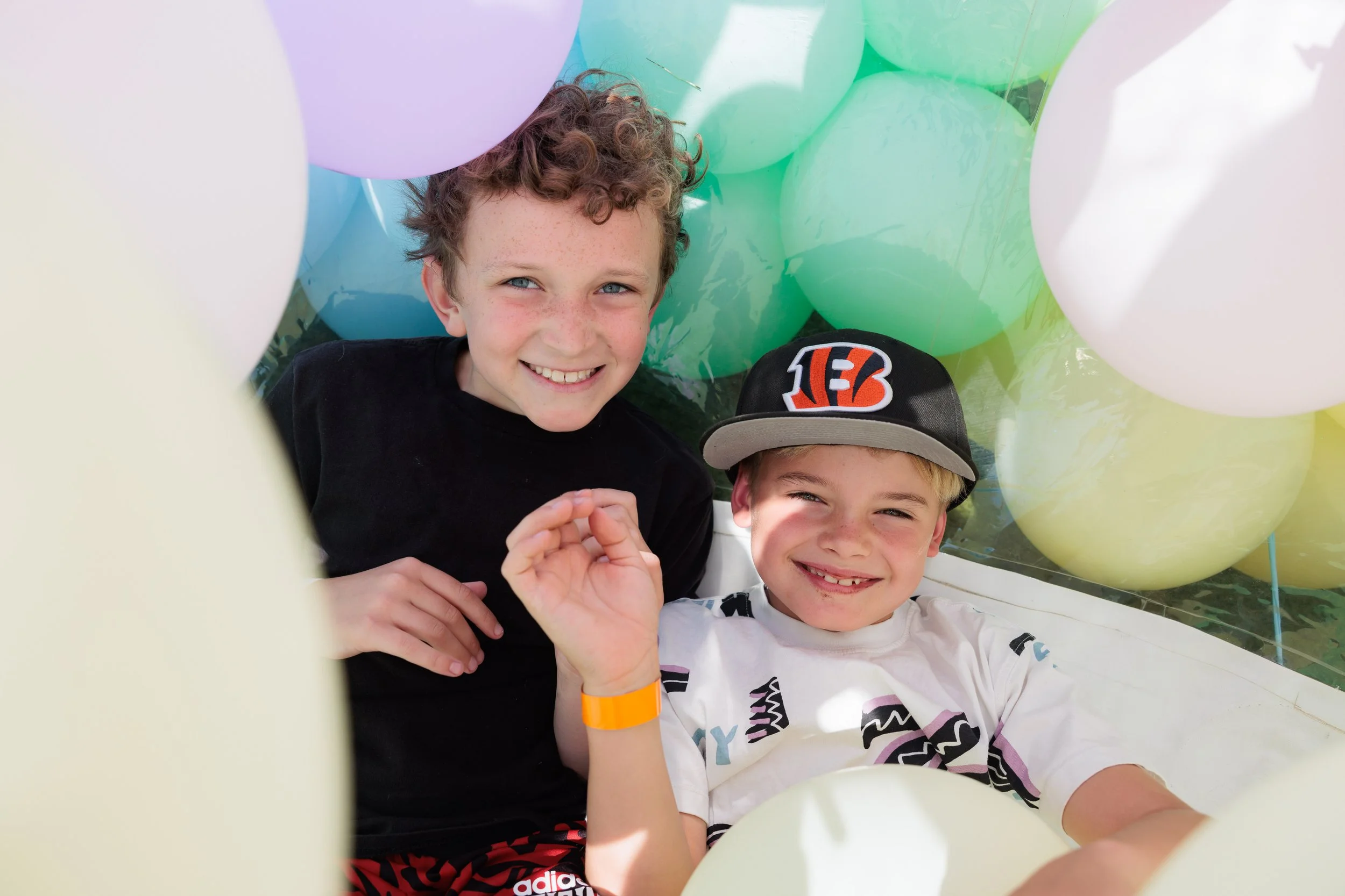 Two boys smiling, surrounded by colorful balloons, one wearing a Cincinnati Bengals cap and the other in a black shirt with a yellow wristband.
