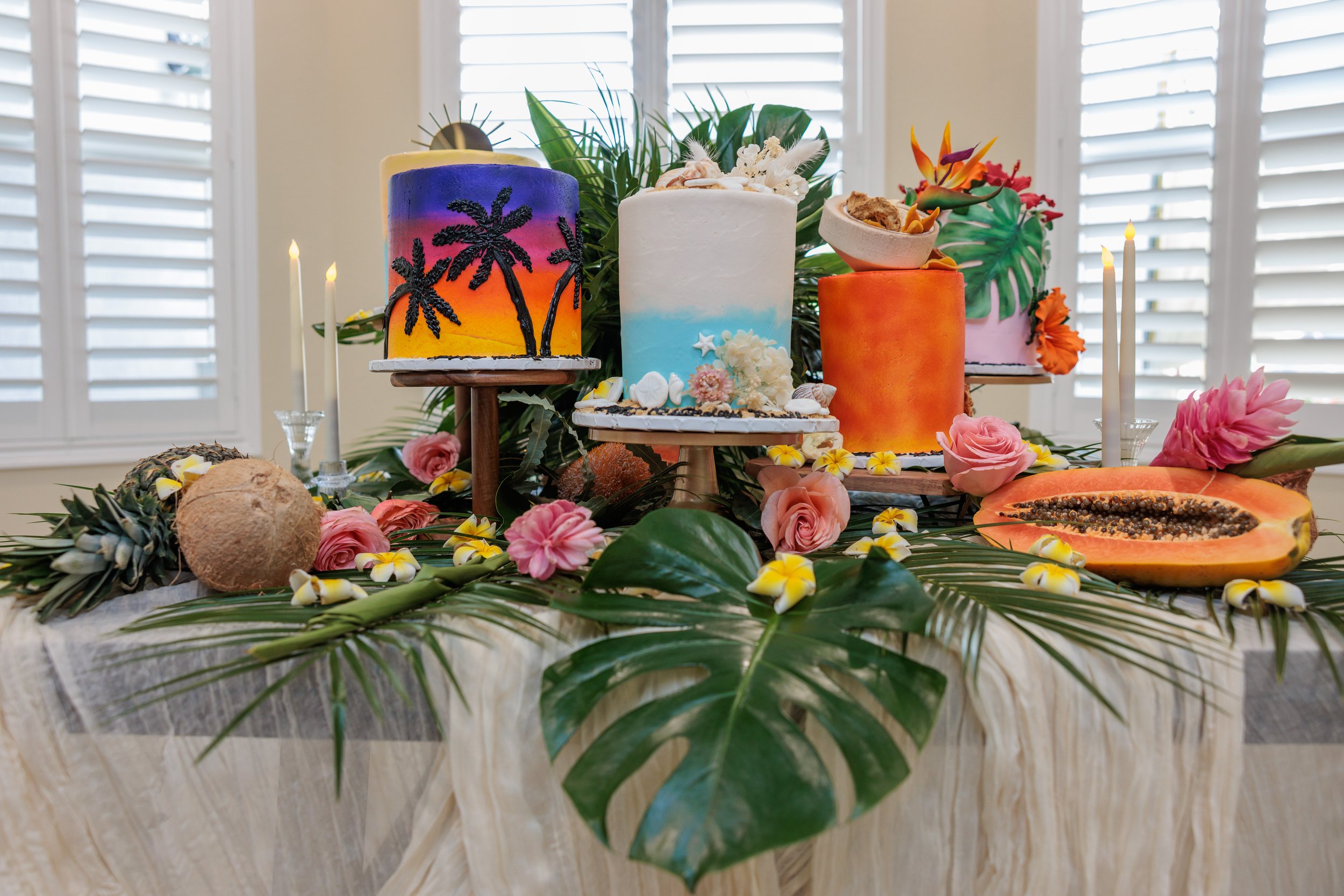 Tropical-themed dessert table with three colorful cakes, tropical fruits, and flowers on a table with tropical leaves, in front of a window with shutters.