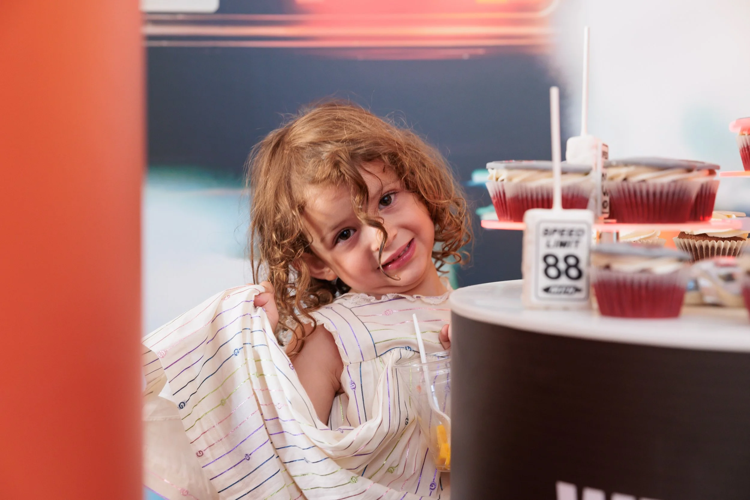 A young girl with curly red hair making a distressed or confused facial expression at a display of cupcakes and a drink.