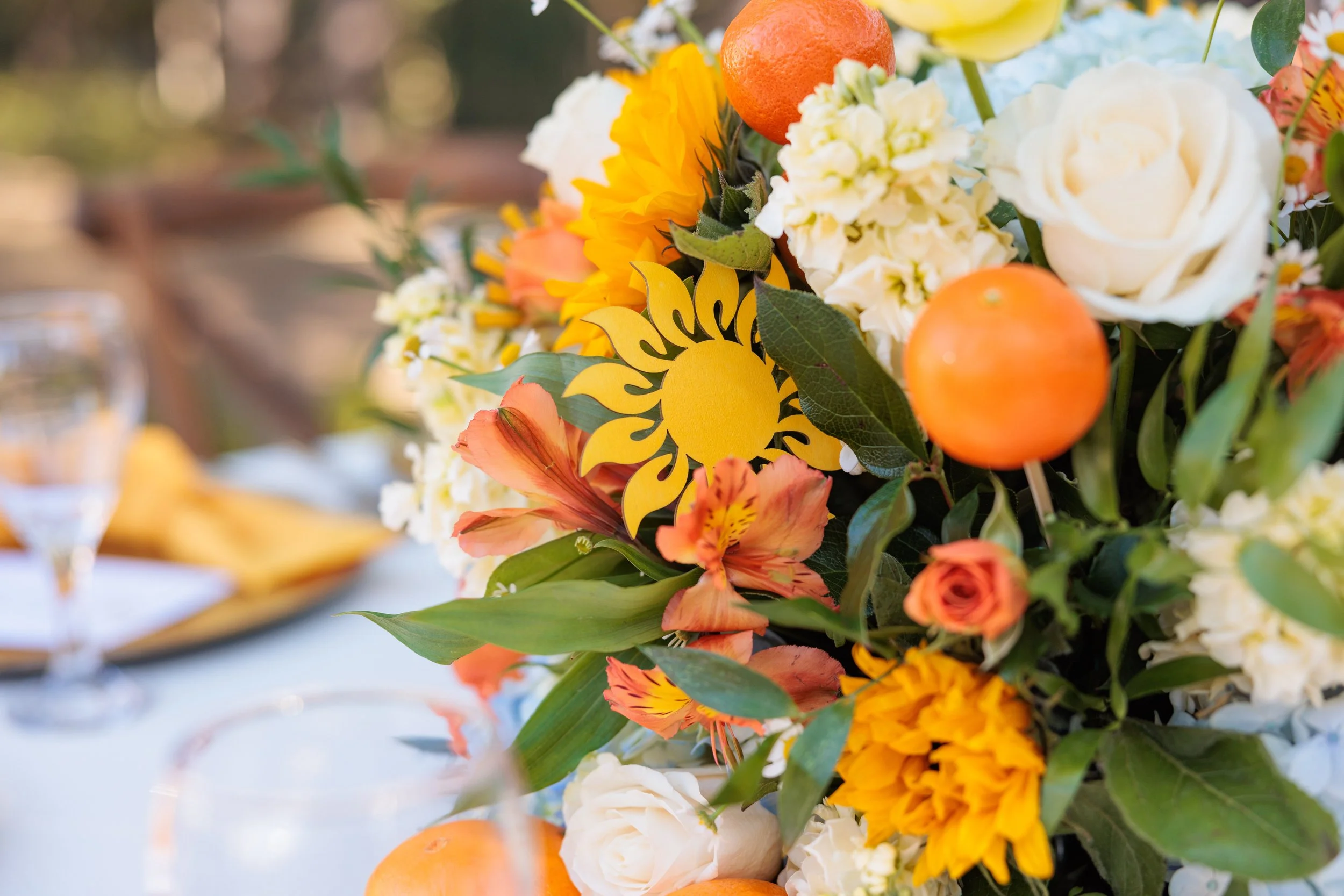 Close-up of a floral arrangement with white roses, orange and yellow flowers, green leaves, and orange decorations, featuring a yellow sun-shaped ornament in the center.