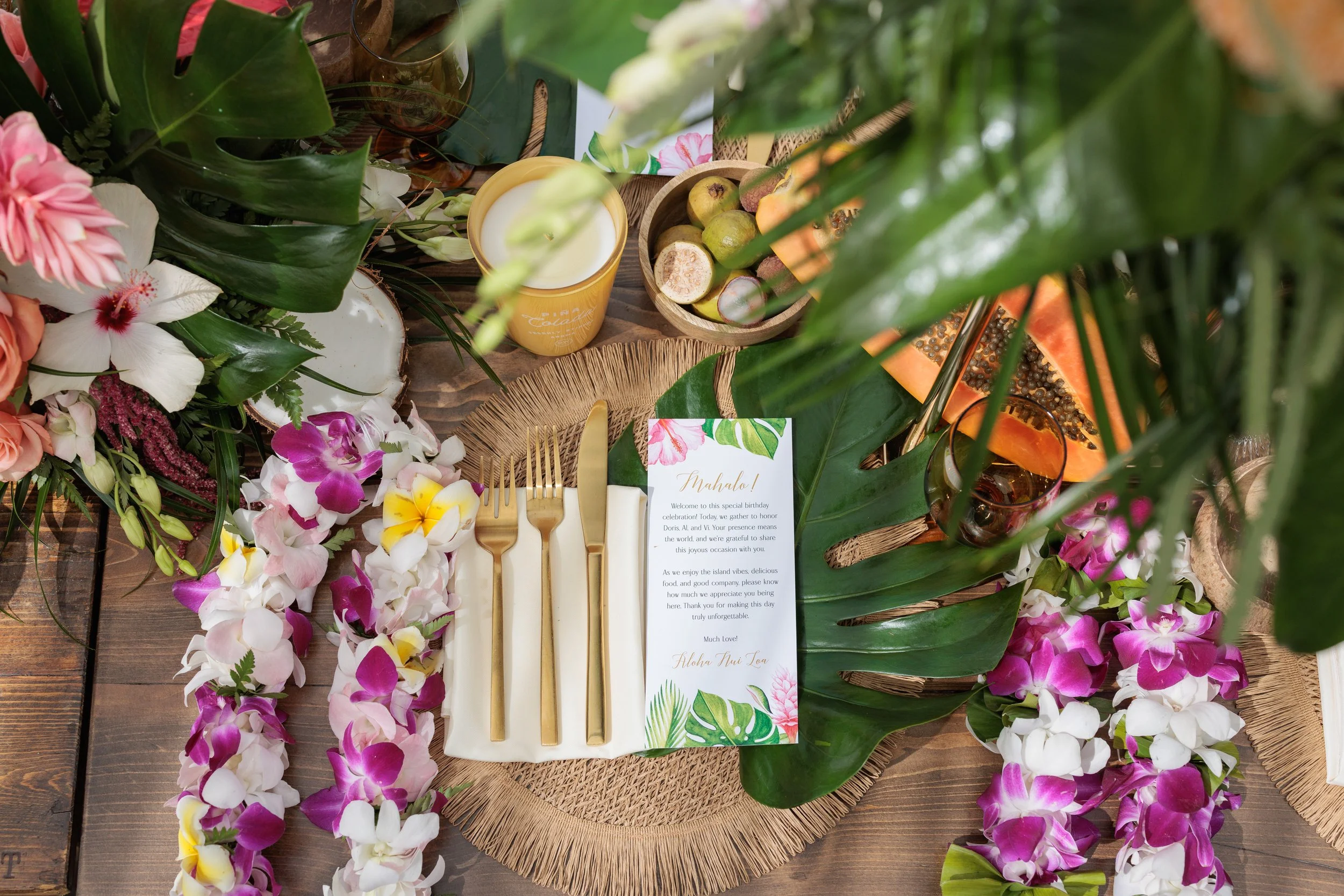 A tropical-themed table setting with pink, white, and purple orchids, green leaves, a card with floral design, gold cutlery, a drink, and a papaya on a rustic wooden table.
