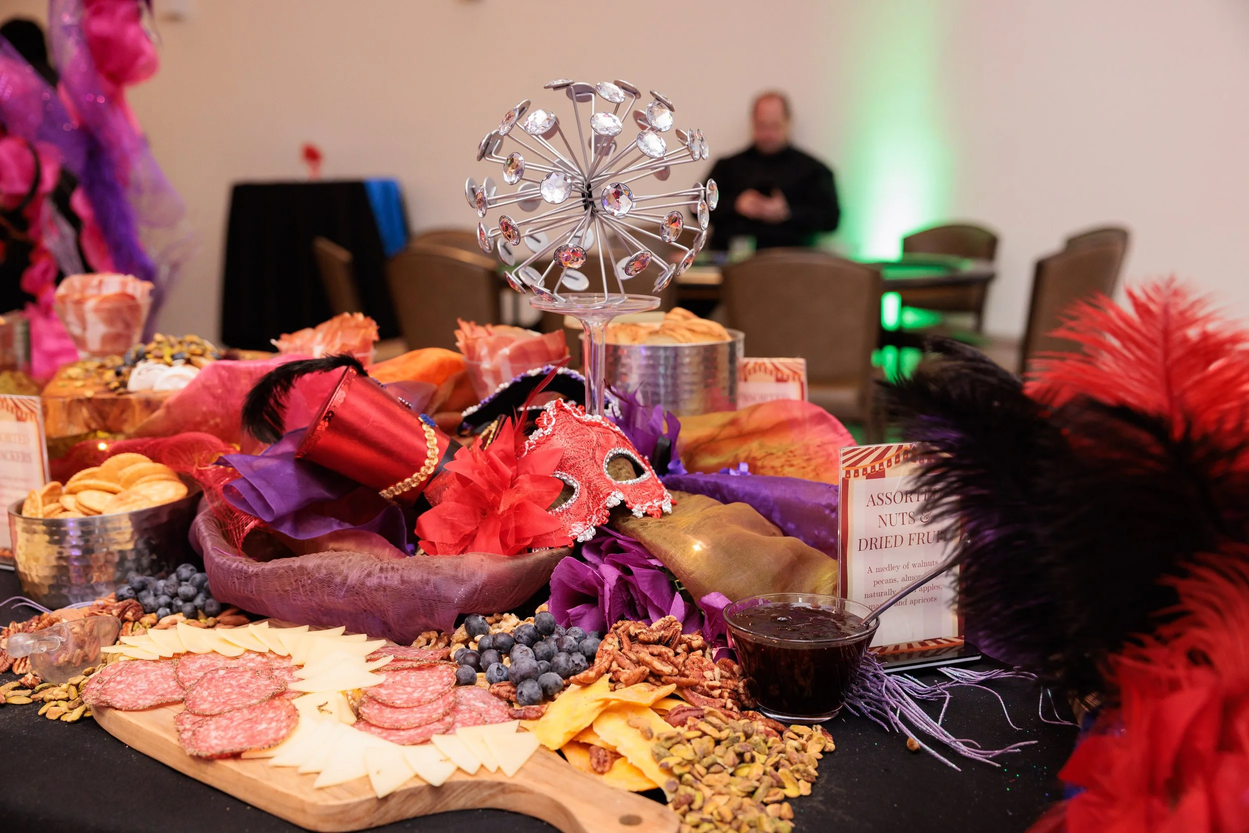 A table decorated with a Mardi Gras theme features masks, feathers, masks, meats, cheeses, nuts, and fruits, with a colorful, vibrant display for a celebration.