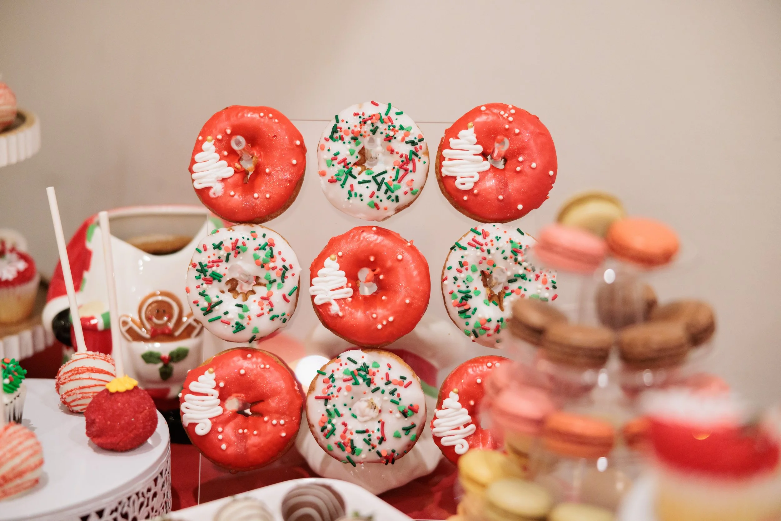 Display of decorated donuts with red and white icing, green and red sprinkles, surrounded by assorted macarons and chocolates for a festive celebration.