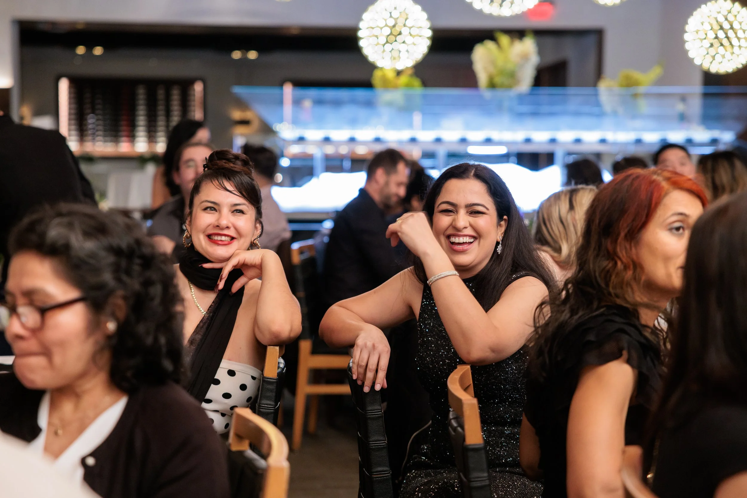 Group of women dressed in formal attire, smiling and laughing at a social event in a well-lit restaurant or banquet hall.