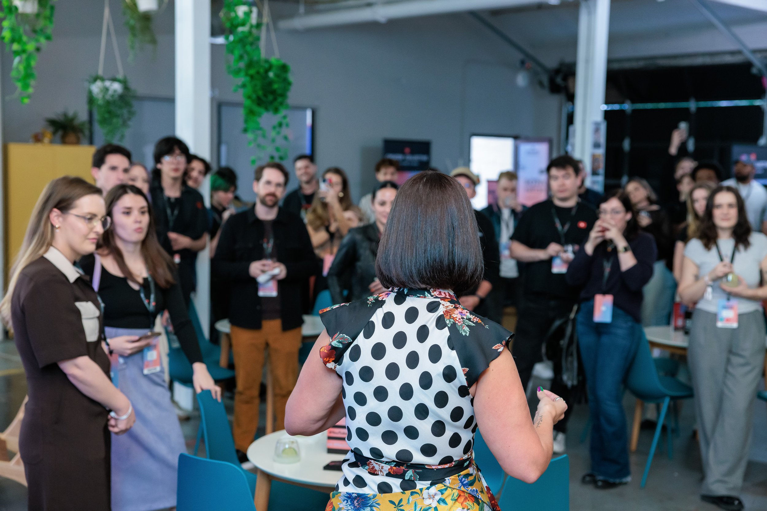 A woman with short dark hair, wearing a black dress with floral print on the shoulders and a polka dot back, is speaking to a group of people at a gathering or conference inside a modern venue with hanging green plants and people wearing name tags.