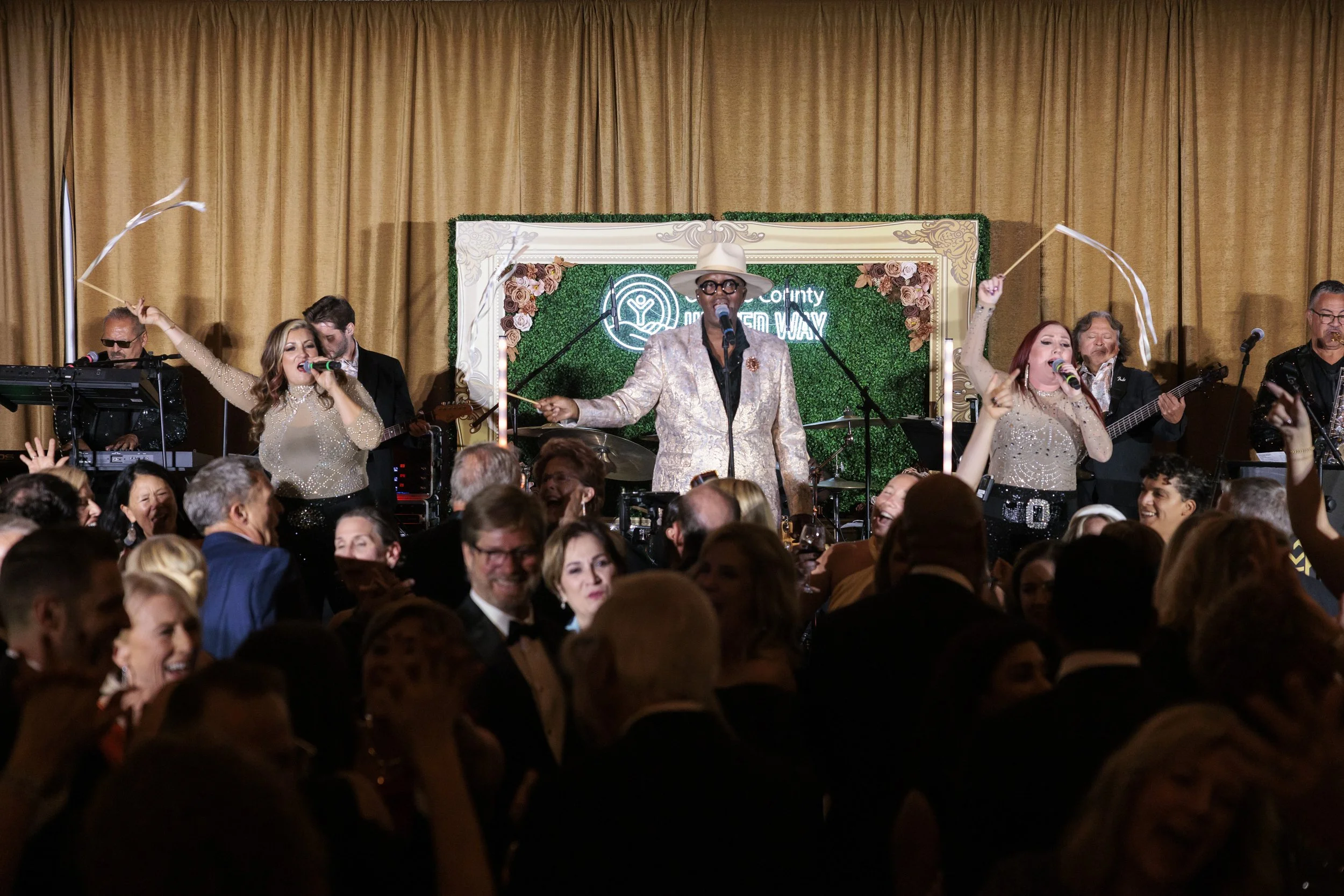 Performer in a glittery suit and wide-brimmed hat singing into a microphone on stage at a lively event, with a band and audience.