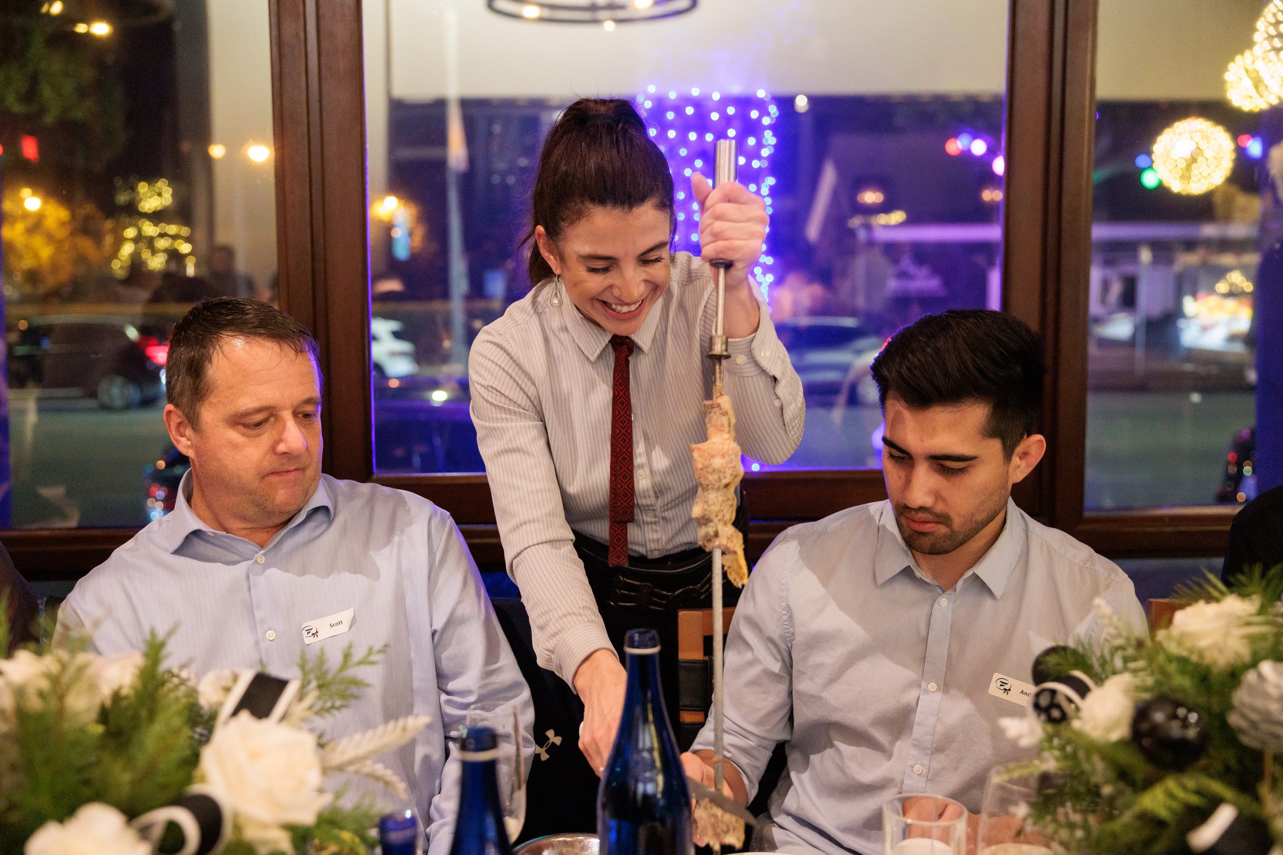A woman serving food using long tongs to two seated men at a dinner table in a restaurant, with festive lights outside the window.