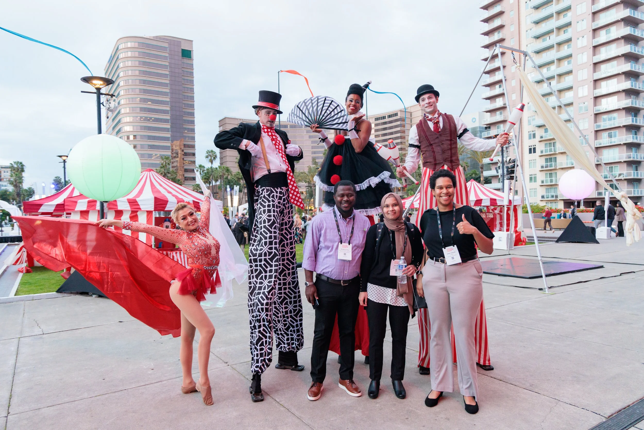 A group of six people posing in front of circus performers on stilts, with tents and high-rise buildings in the background. The performers are dressed as clowns and acrobats, with colorful costumes and props, while the group appears to enjoy the even