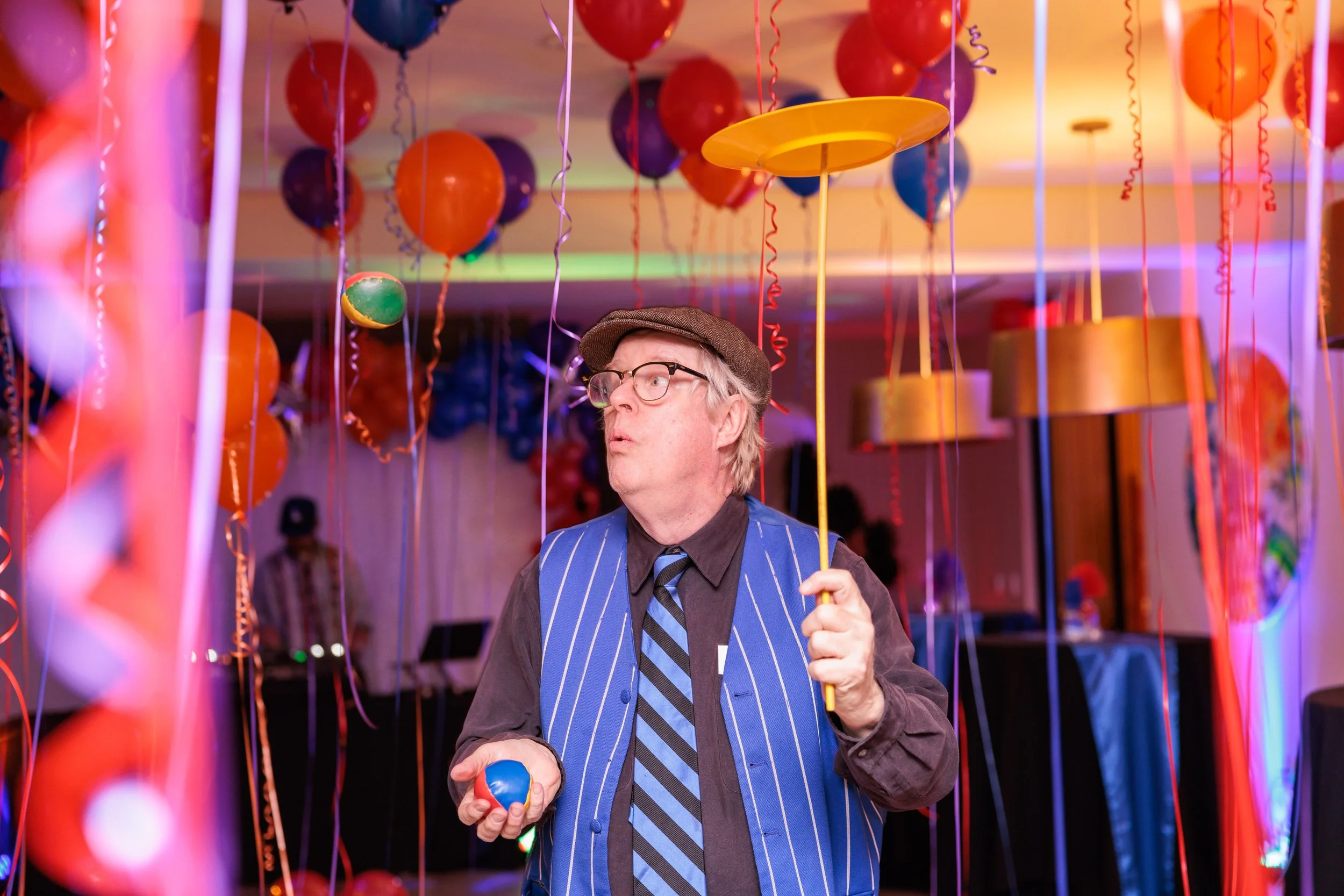 A man dressed in a dark shirt, striped vest, and tie, wearing a cap and glasses, stands at a party decorated with balloons and streamers, holding a juggling ball in one hand and a yellow umbrella in the other, with colorful lights illuminating the sc
