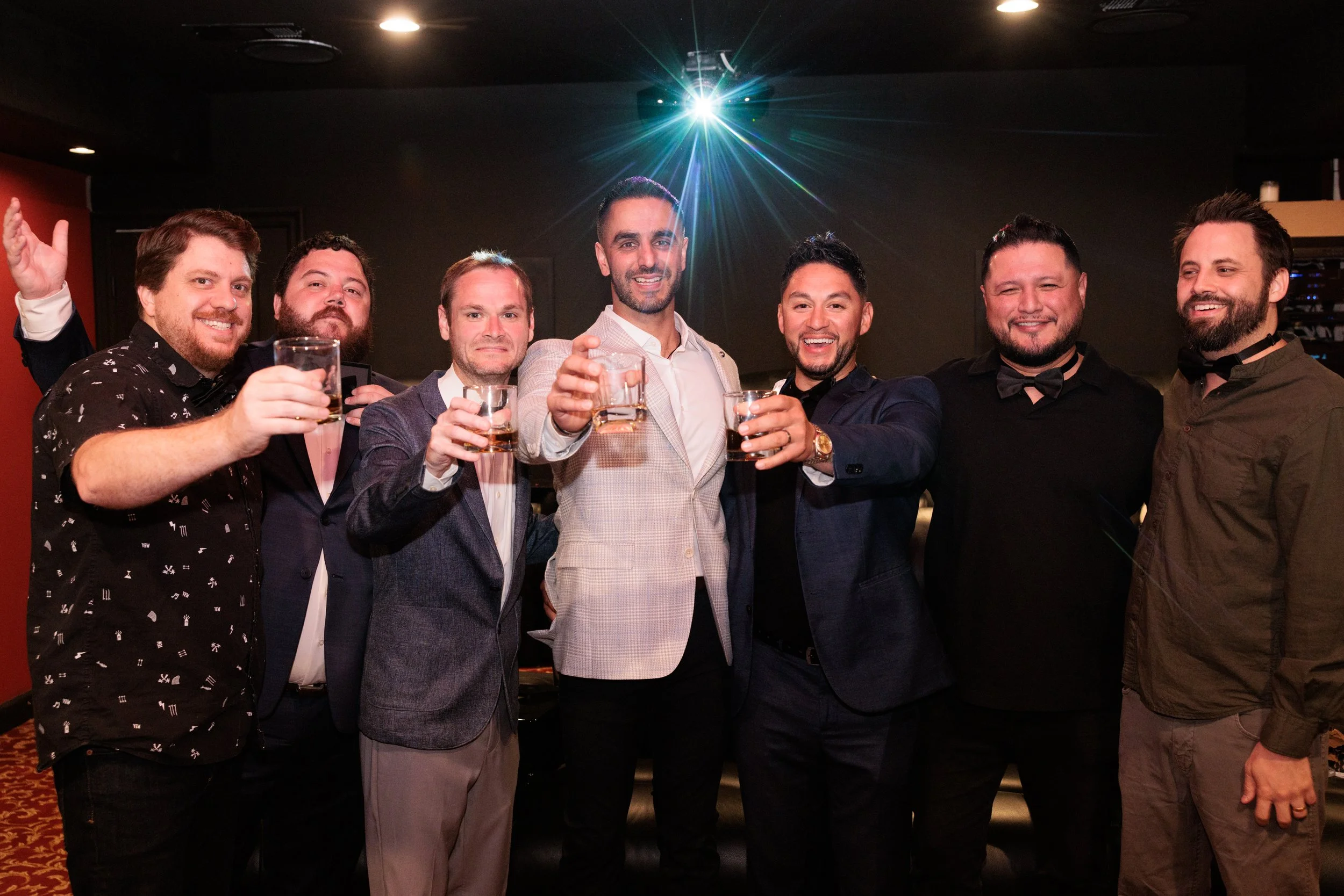 A group of eight men in formal and semi-formal attire, holding glasses of drinks, smiling and celebrating together at a party or social gathering in a dimly lit room with a disco ball or bright light above.