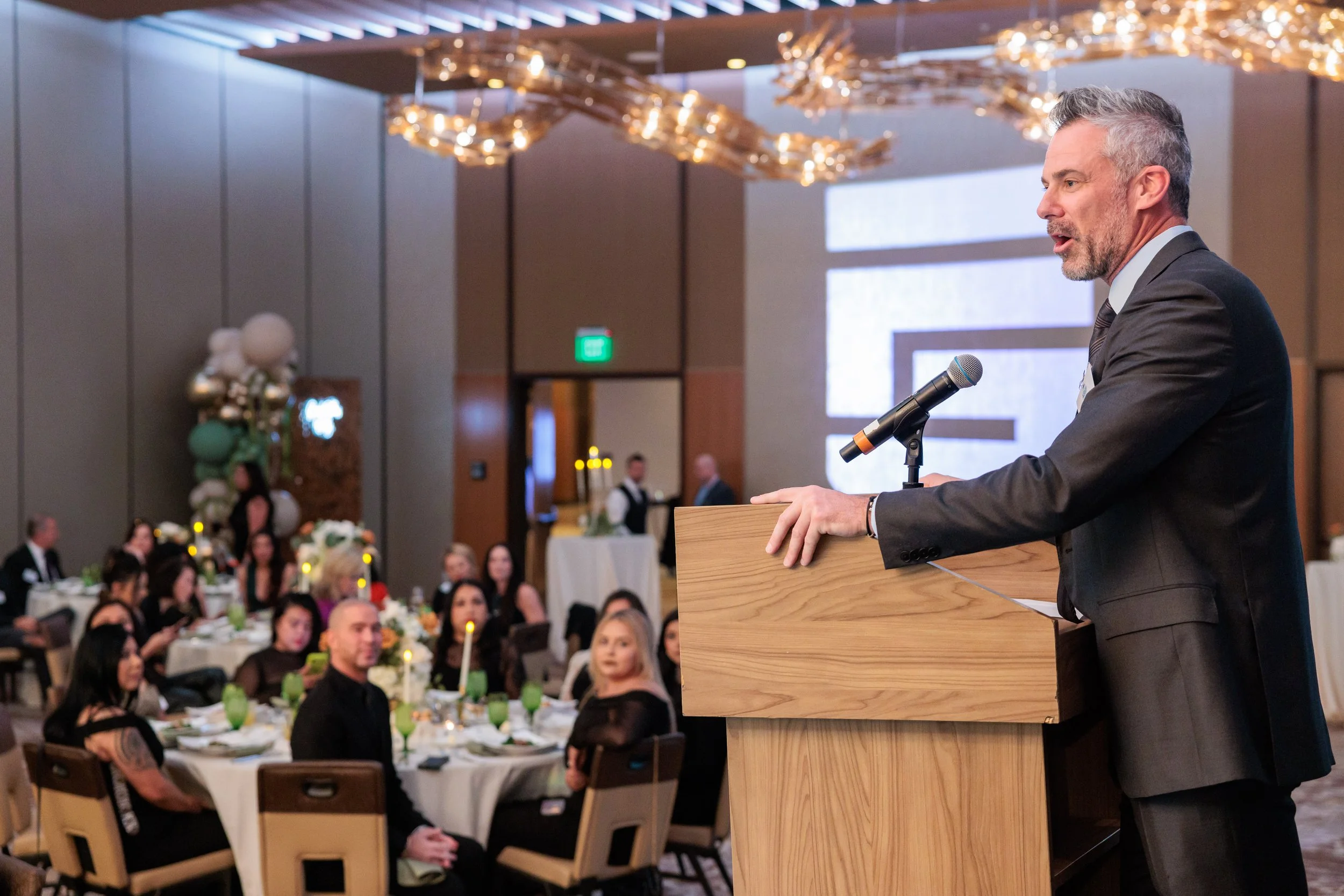 A man in a suit speaking at a podium with a microphone during an event or banquet. The audience is seated at round tables decorated with candles and floral centerpieces, and the room has modern lighting and decor.